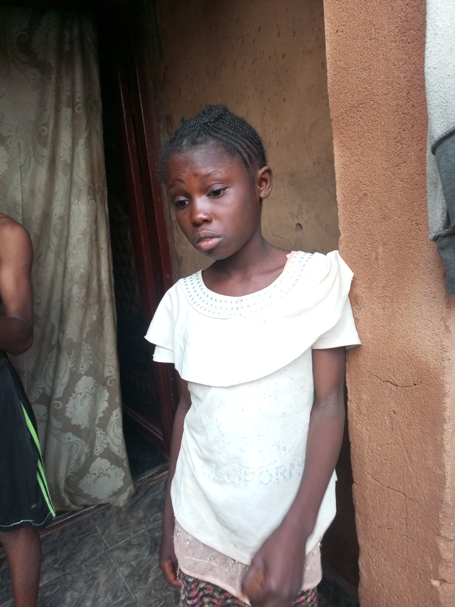 A young Black girl stands near a doorway, leaning against a textured, reddish-brown wall. She has dark skin and her hair is styled in braids. She is wearing a white t-shirt with a ruffled neckline and some small, shimmering embellishments that appear to form text on the front. Underneath the t-shirt, a pink lace hem is visible, along with a patterned skirt. To her left, a curtain with an ornate pattern hangs, and part of another person's arm and legs, wearing black shorts with a green stripe, is visible. The ground appears to be tiled. The lighting suggests it is daytime, possibly outdoors. The girl's expression is somber, with her gaze directed downwards. The overall scene suggests a casual, everyday moment in Gombe, Nigeria.