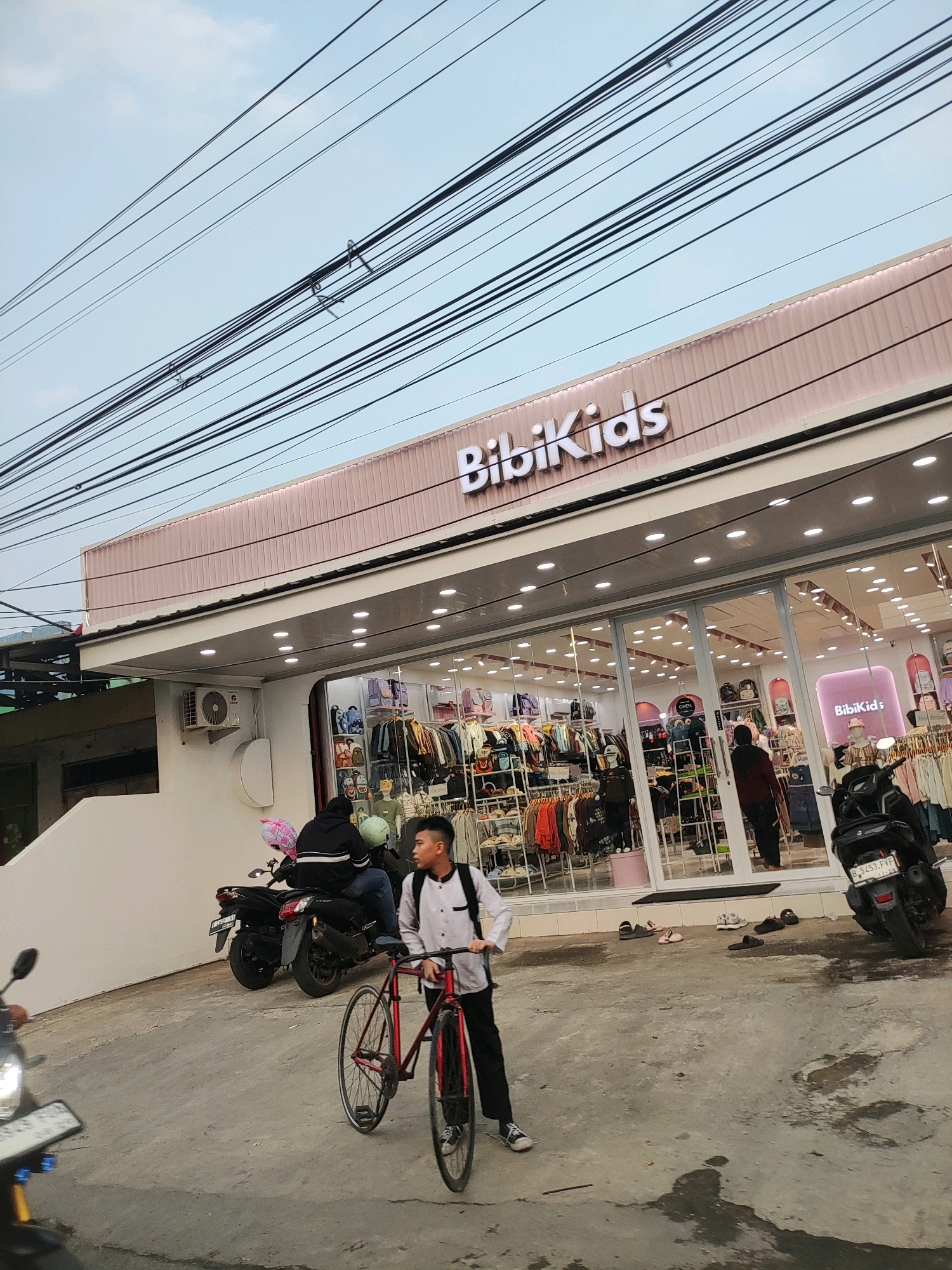 In the city of Wanajaya, Indonesia, a street scene is captured featuring a storefront for "BibiKids," a children's clothing store. The store has large glass windows displaying racks of colorful children's apparel and accessories. Above the entrance, the "BibiKids" logo is prominently displayed. A young male, dressed in a white collared shirt and black pants, walks towards the right of the frame, pushing a red bicycle. Behind him, a dark-colored motorcycle is parked, with two individuals seated on it, one wearing a black hoodie and the other a light-colored helmet. Another motorcycle, partially visible in the foreground on the left, has its headlight on. The street surface appears wet in patches, suggesting recent rain. Overhead, a complex network of power lines crisscrosses the sky, characteristic of urban infrastructure. The sky is overcast.
