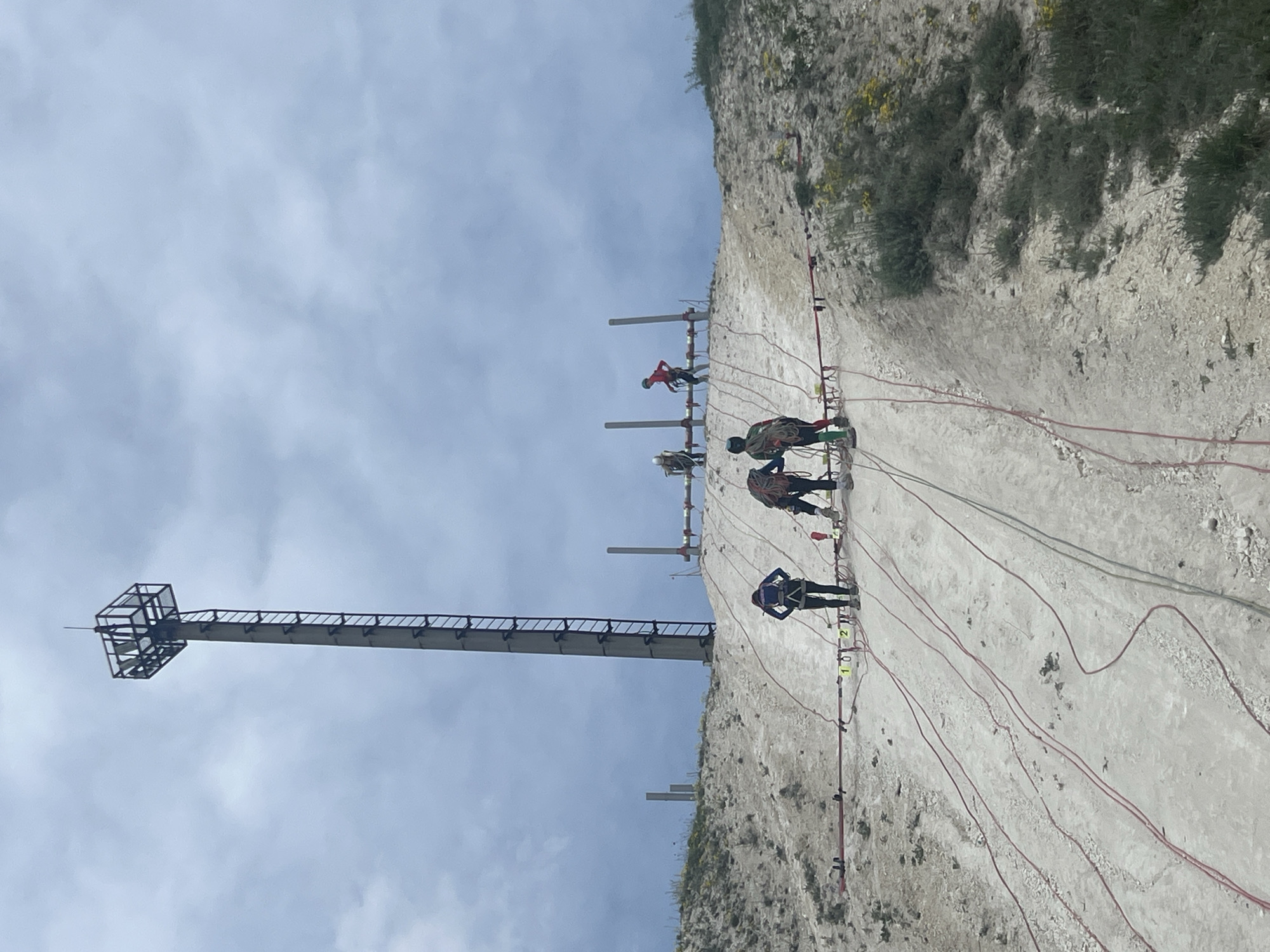 The image depicts a rock climbing facility in Khvalynsk, Russia. A tall, metal structure with a ladder-like extension stands to the left of the climbing wall. Four individuals are actively climbing the wall, secured by ropes and harnesses. The climbing wall is textured and has vegetation growing on top. The sky above is partially cloudy.