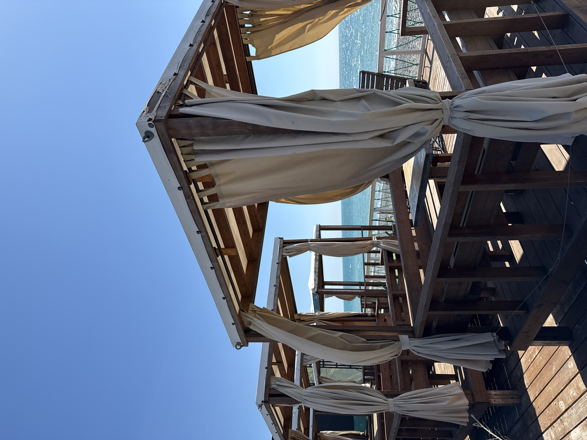 The image presents a serene view of a series of wooden beach cabanas or pergolas extending towards the sea under a clear blue sky. The structures are constructed from dark brown wood, forming a lattice framework. Light beige or off-white fabric curtains are tied back to the wooden posts on many of the cabanas, suggesting they are prepared for use or have been tidied. Some drapes are partially unfurled, with gentle folds.
Beyond the wooden structures, the bright blue sea shimmers in the sunlight, indicating a sunny day, likely morning or early afternoon given the high angle of the sun and distinct shadows cast by the wooden beams on the decking. A pebbly shoreline is visible where the wooden structures meet the water. The sky is a uniform, vibrant blue, without clouds. The overall scene suggests a calm and inviting coastal resort environment. No people are visible, and there is no discernible text in the image. The setting is consistent with a beachside leisure area in a location like Sochi, Russia.