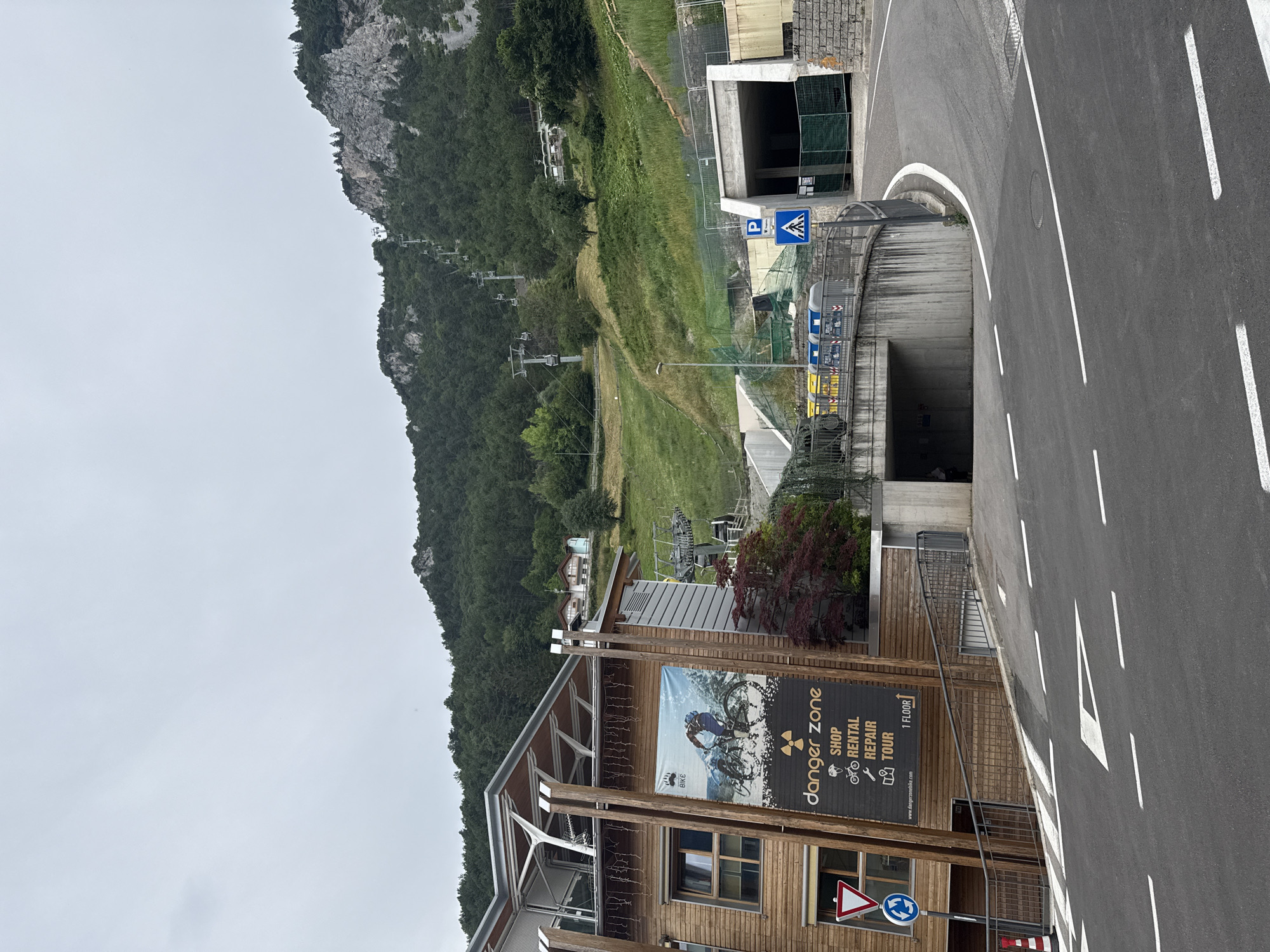 The image depicts the entrance to a tunnel and a ski lift station in the mountainous terrain of Molveno, Italy. A paved road curves into the frame, leading towards the tunnel entrance. On the left, a wooden building with a contemporary design features a large banner advertising "Danger Zone," with text indicating "Shop," "Rental," "Repair," and "Tour." A sign with a downward-pointing arrow and "1 FLOOR" is also visible on the banner. The building has several windows and is supported by wooden beams.
Above the building and extending up the mountainside, a ski lift system with multiple cabins is visible. The mountain slope is covered in lush green trees and grass, with a clear, overcast sky overhead. Several smaller buildings, possibly houses, are nestled on the hillside. On the road, a pedestrian crossing sign and a parking sign are posted near the tunnel entrance. Construction barriers and fencing are present around the tunnel area, suggesting ongoing work. The overall scene is a blend of urban infrastructure, recreational facilities, and the natural beauty of the Italian Alps.
