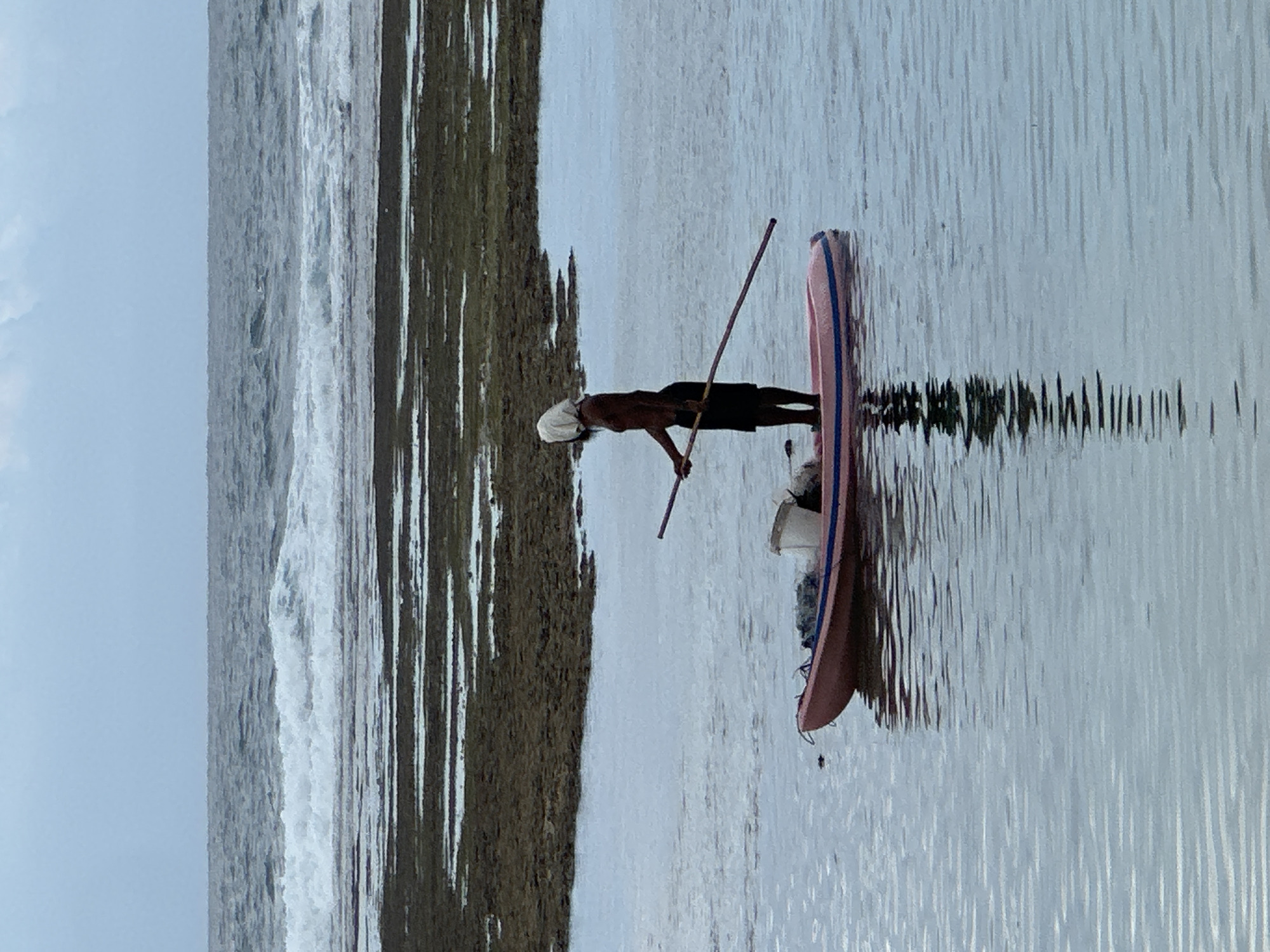 A lone individual, clad in black shorts and a white head covering, stands on a pink inflatable boat in the shallow waters of Kuta Selatan, Indonesia. They hold a long pole, likely for navigation or fishing, with the tip extended towards the right side of the frame. A white bucket and fishing net are visible within the boat. The water reflects the sky and the boat, creating ripples that distort the scene. In the background, the ocean churns with waves crashing against a dark, rocky shore, under a cloudy sky.