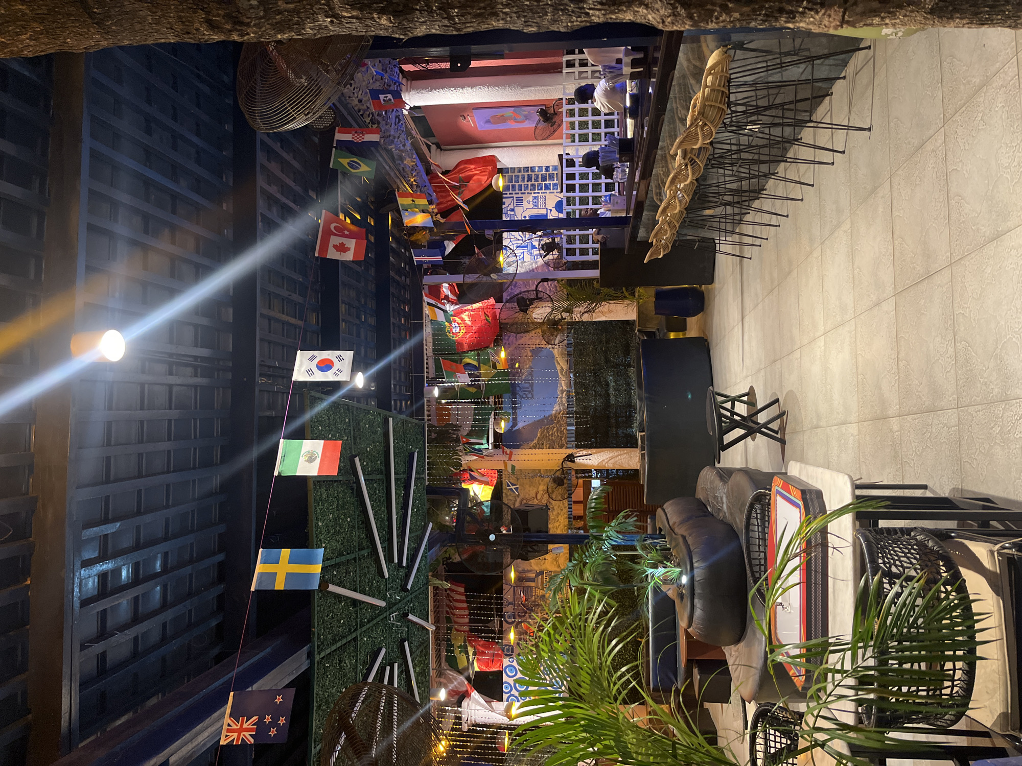 The image depicts an outdoor patio area of a restaurant or bar in Accra, Ghana, at night. The space is decorated with numerous national flags strung across the ceiling, creating a festive atmosphere. String lights and ceiling lights illuminate the area. In the foreground, there are tables with chairs, some of which are woven wicker. A large, dark, modular sofa with cushions is a prominent seating arrangement. Plants in pots add greenery to the scene. In the background, a bar area is visible with stools and patrons seated at the counter, illuminated by soft lighting. There is a textured wall and pillars, with some decorative elements and paintings. The flooring appears to be tiled.