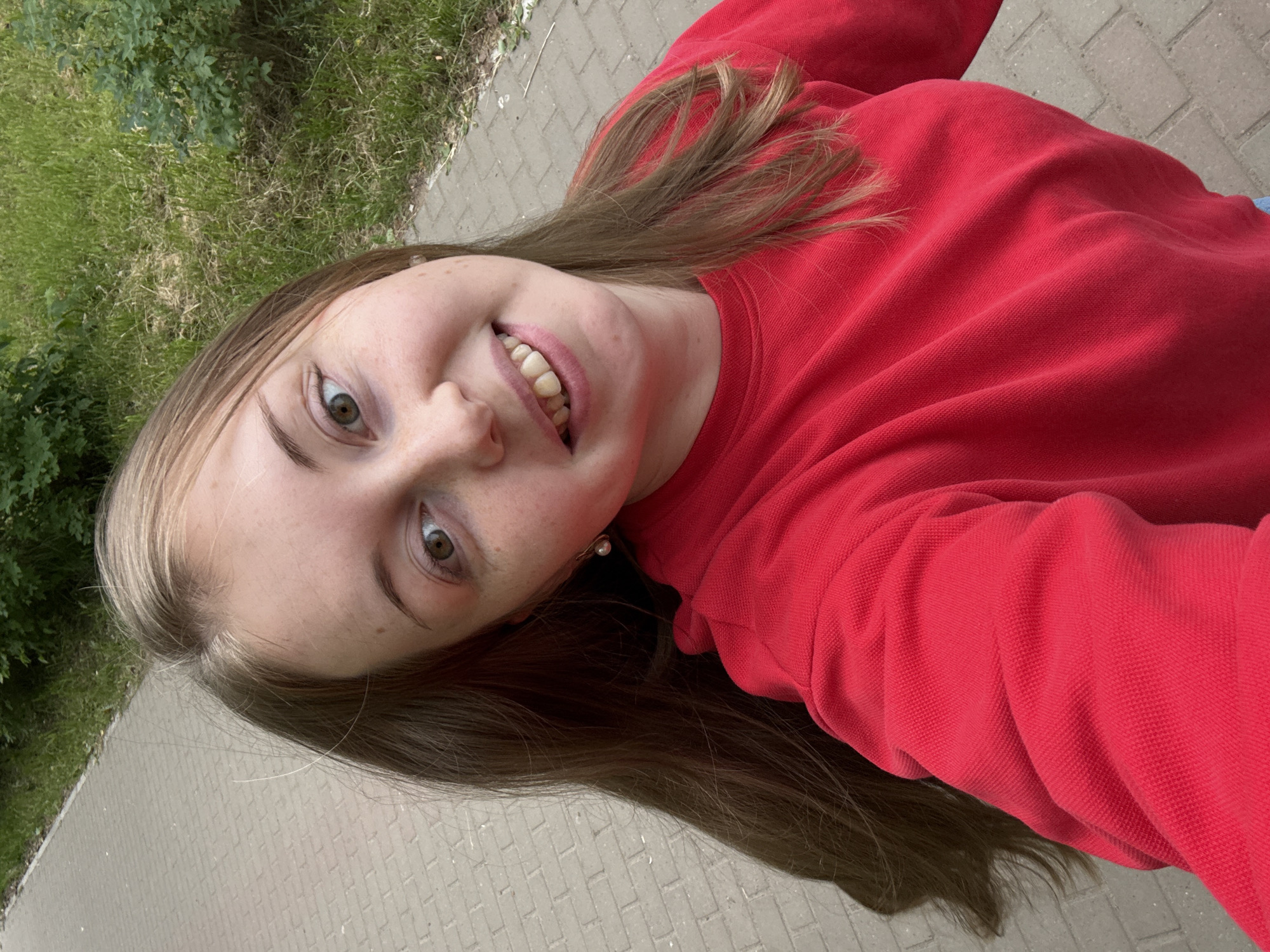 The media file is a selfie taken outdoors, from a low angle, with the subject's head tilted to the left. The subject appears to be a young person with long, brown hair, a red sweater, and freckles across their nose and cheeks. They are smiling, showing their teeth, and looking directly at the camera. A small pearl earring is visible on their left earlobe.
The background consists of a paved walkway made of rectangular grey bricks, bordered by a strip of green grass and some bushes. The lighting suggests it is daytime, and the overall atmosphere is casual and cheerful. No text is visible in the image. The orientation of the image suggests it was taken by someone holding the camera themselves.