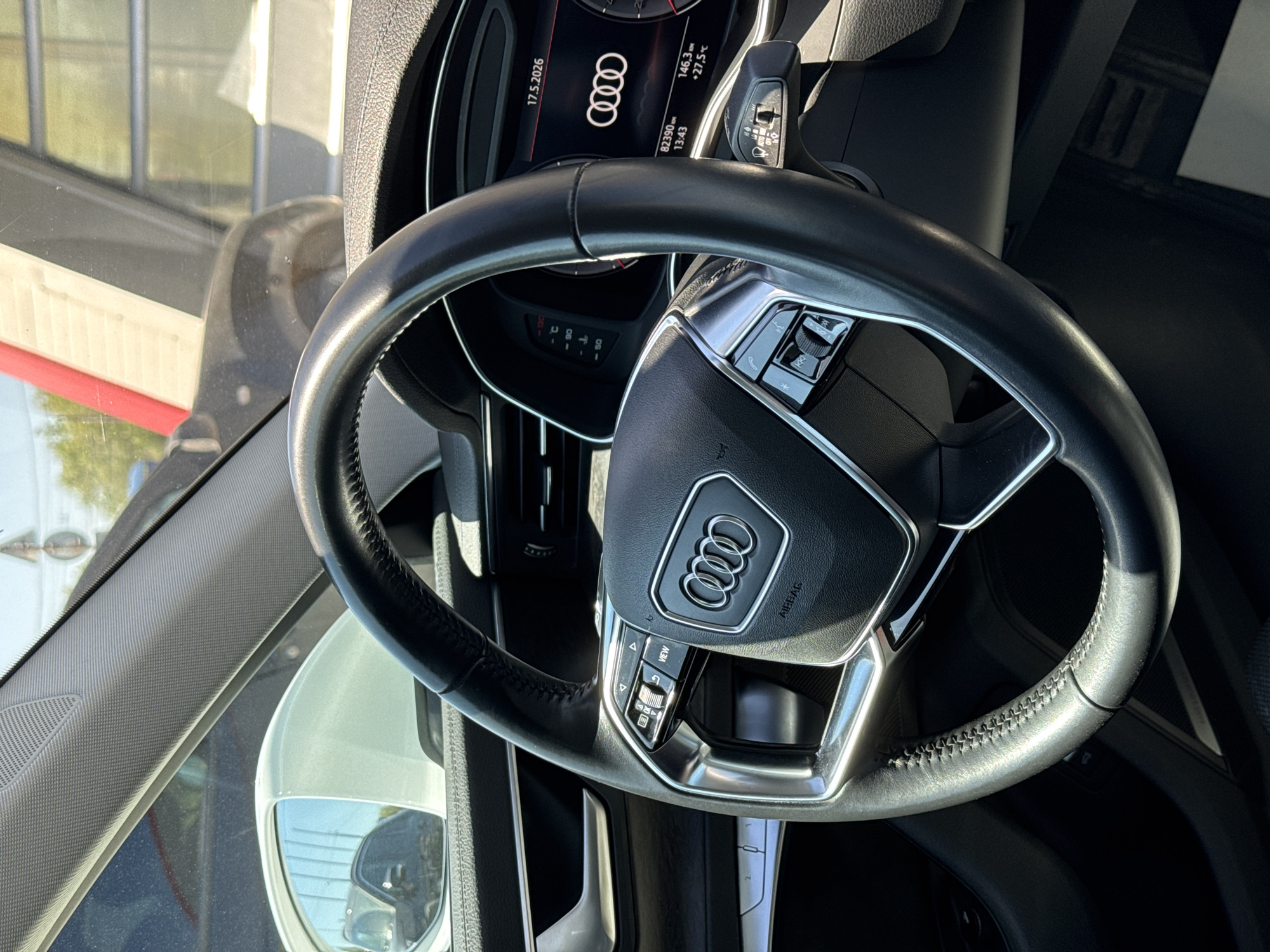 The image shows the interior of an Audi vehicle, specifically the steering wheel and dashboard area. The steering wheel is black leather with silver trim, featuring the Audi logo in the center. The dashboard displays information such as the date (17.5.2026), the Audi logo, and vehicle statistics (e.g., 146.3 km, 27.5°C, 82390 km). The environment suggests the vehicle is stationary, as indicated by the clear view through the windshield, showing the exterior surroundings. The location, based on the provided details, is an unidentified area within the city of Unknown, Unknown.
