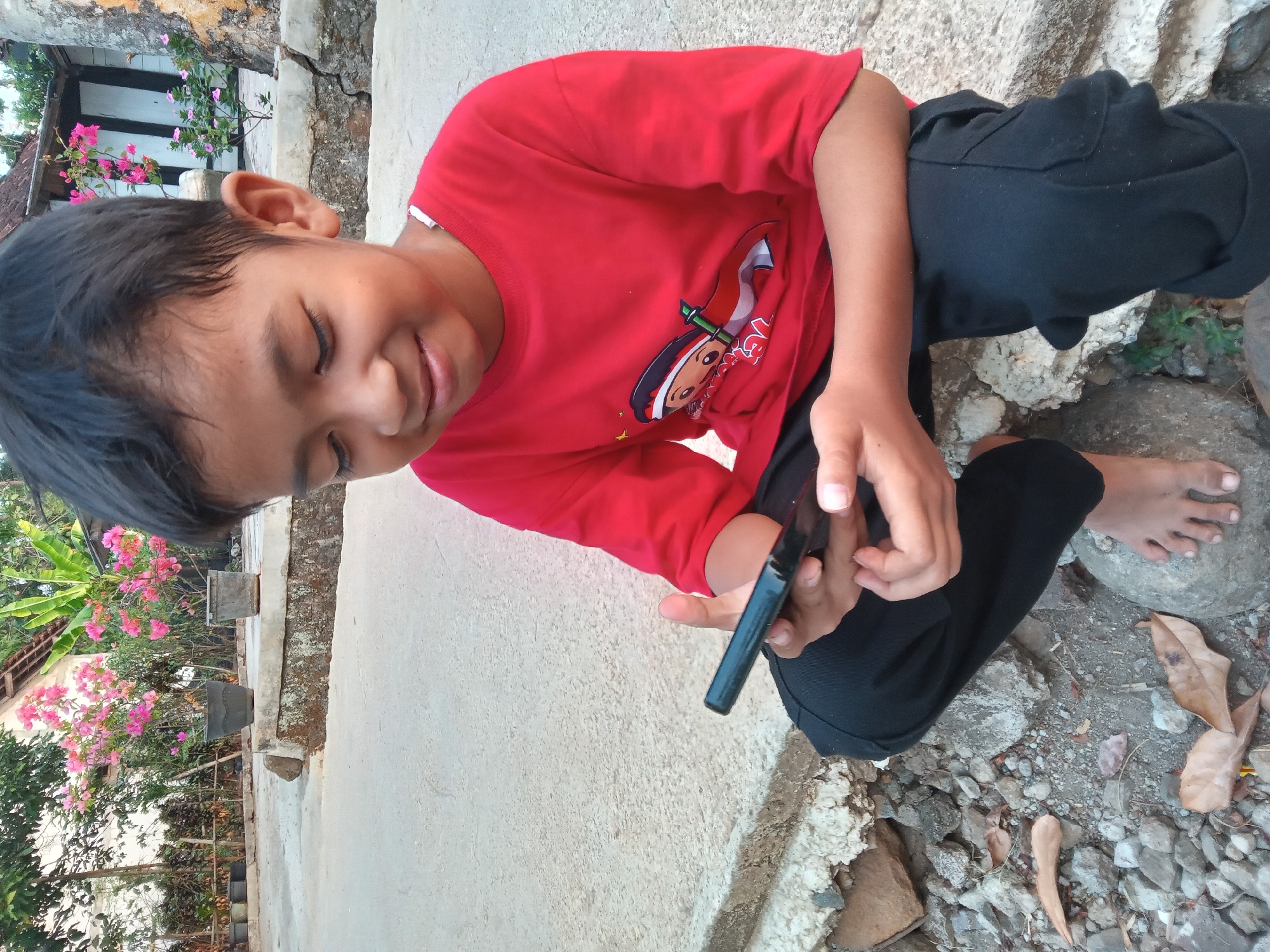 A young boy wearing a red t-shirt with a graphic of a cartoon character holding an Indonesian flag and black pants is sitting outdoors on a concrete surface, looking down at a black smartphone he is holding in his hands. He has dark hair and a smiling expression. The background features potted pink flowers, green foliage, and part of a building. The ground is a mix of concrete and rough stones, with some dry leaves scattered around. The scene appears to be in an outdoor, possibly residential, setting in Kedunggudel, Indonesia.