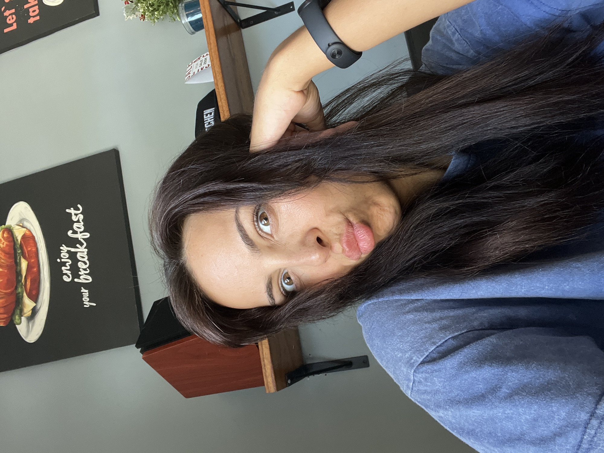 A woman with long, dark hair and brown eyes is photographed indoors. She is wearing a black smartwatch on her left wrist and a blue shirt. Her right hand is near her head. Behind her are wall decorations, including a framed breakfast image. The location is potentially Kuta Selatan, Indonesia, but there are no visual elements to confirm the location definitively.