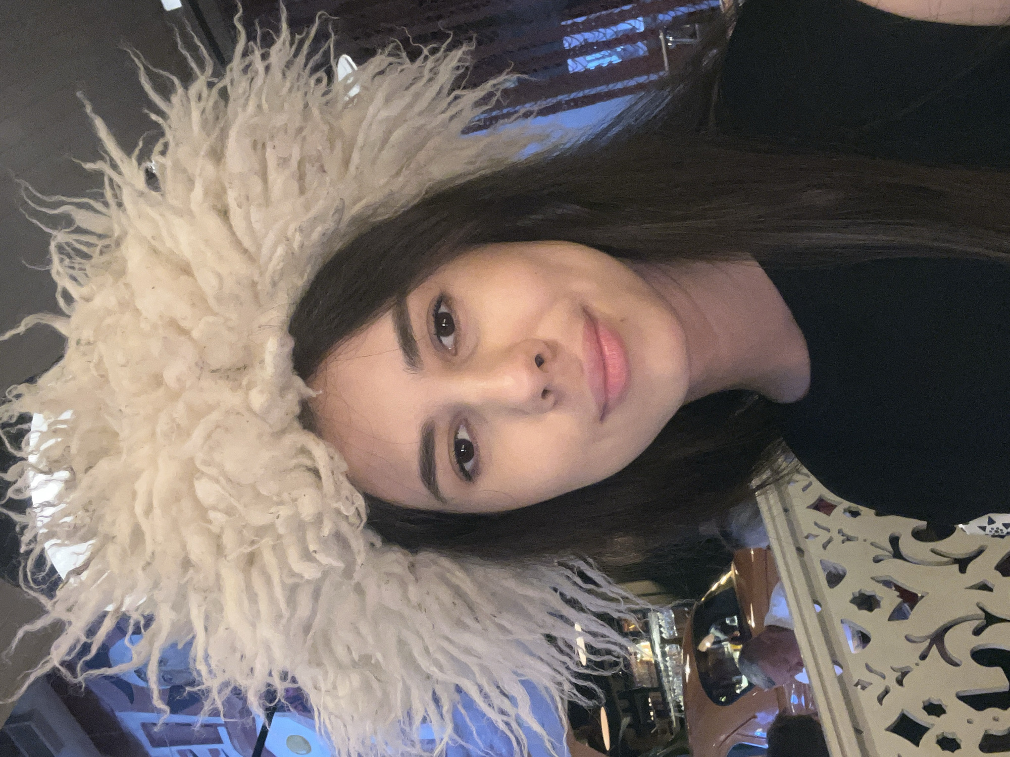 The image shows a close-up, sideways portrait of a young woman wearing a fluffy, cream-colored hat or hood. She has long, dark hair and is looking towards the camera with a slight smile. Her eyes are brown, and she appears to be wearing subtle makeup. The background is blurred, but it suggests an indoor setting with what looks like decorative elements, possibly a patterned screen or railing to the right. The lighting seems artificial, and the overall mood is casual and possibly playful due to her expression and the unique headwear.