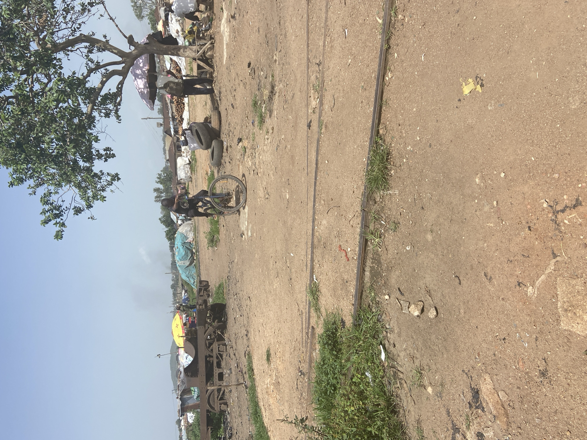 The image depicts an outdoor scene at what appears to be a market or informal settlement. Several individuals are present, some standing near stalls or structures covered with tarpaulins. One person is seated on a bicycle. Scattered items include tires, what seem to be goods for sale, and debris on the ground. A large tree with green foliage is visible on the left side of the frame. The ground is dusty and uneven, with what looks like disused train tracks running through the middle of the scene. In the background, more structures and people are visible, suggesting a populated area. The sky is clear and bright, indicating daytime and likely fair weather. The overall impression is one of a busy, potentially impoverished, community setting. There is no visible text in the image.