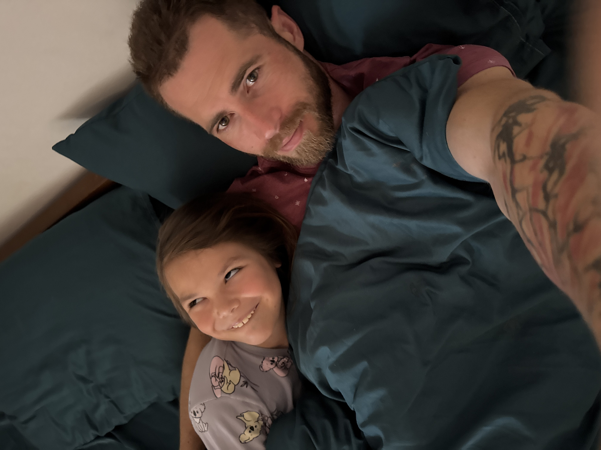 A Caucasian man with a beard and arm tattoos is lying in a bed with dark green sheets and a matching pillow alongside a Caucasian child with short brown hair, wearing a koala-themed shirt. They appear to be taking a selfie. The location is not specified and there is no indication the picture was taken in Budva, Montenegro.