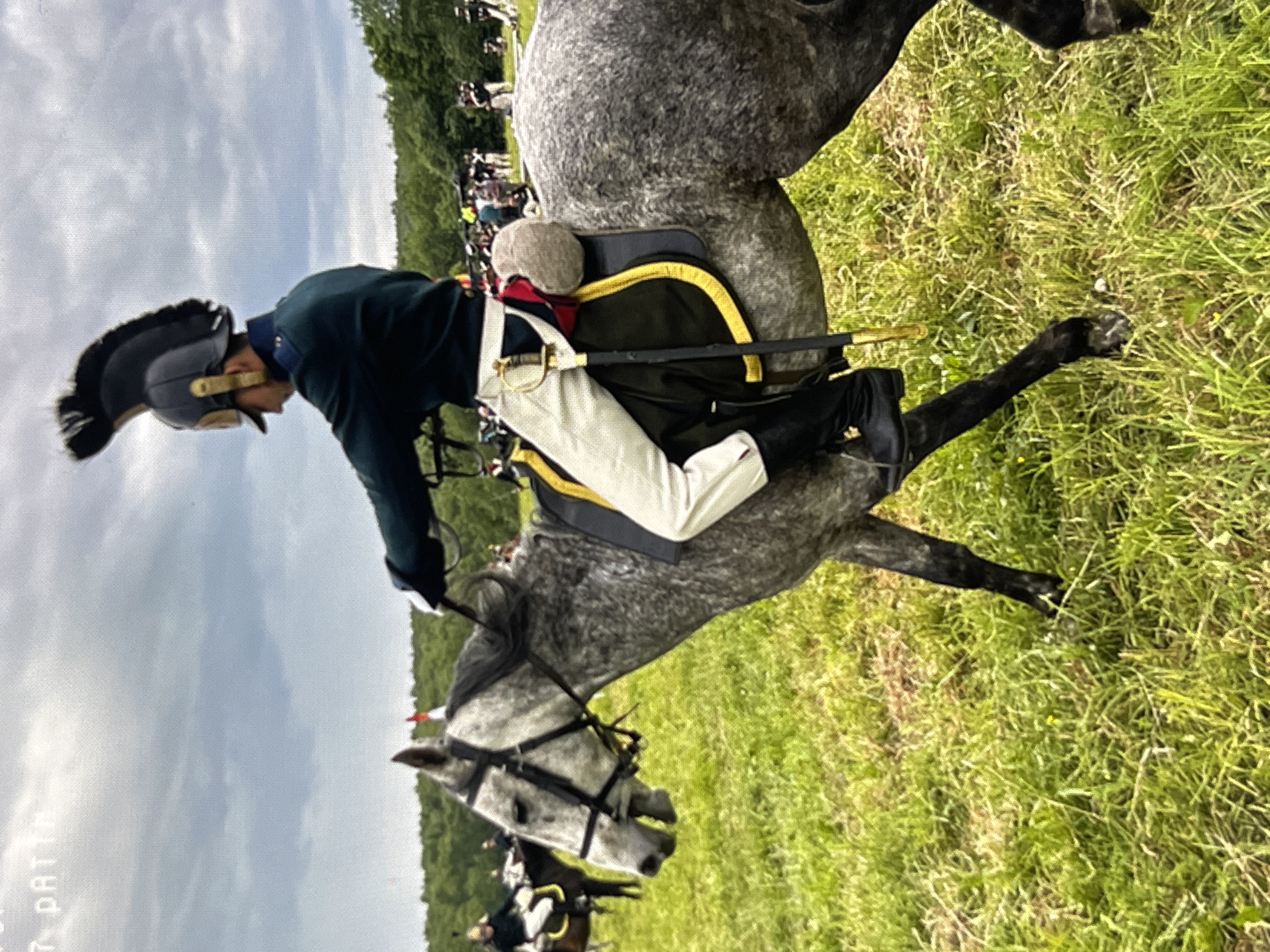 The image shows a man in historical military attire riding a grey horse through a grassy field. The man is wearing a dark green jacket, white breeches, tall black boots, and a helmet with a prominent plume. He has a sword at his side. The horse is equipped with a saddle and bridle. In the background, other individuals on horseback and standing figures are visible, suggesting a reenactment or historical event. The sky is cloudy. There is no discernible information in the image that would place the scene in Moscow, Russia.