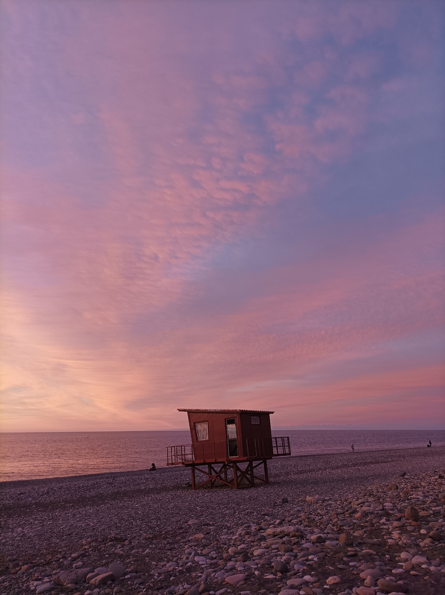 A beach in Unknown, Unknown is seen in this image. The scene shows a small lifeguard station on the rocky shoreline, with the sun appearing to be setting, creating a pink and purple sky. Several people are on the beach.