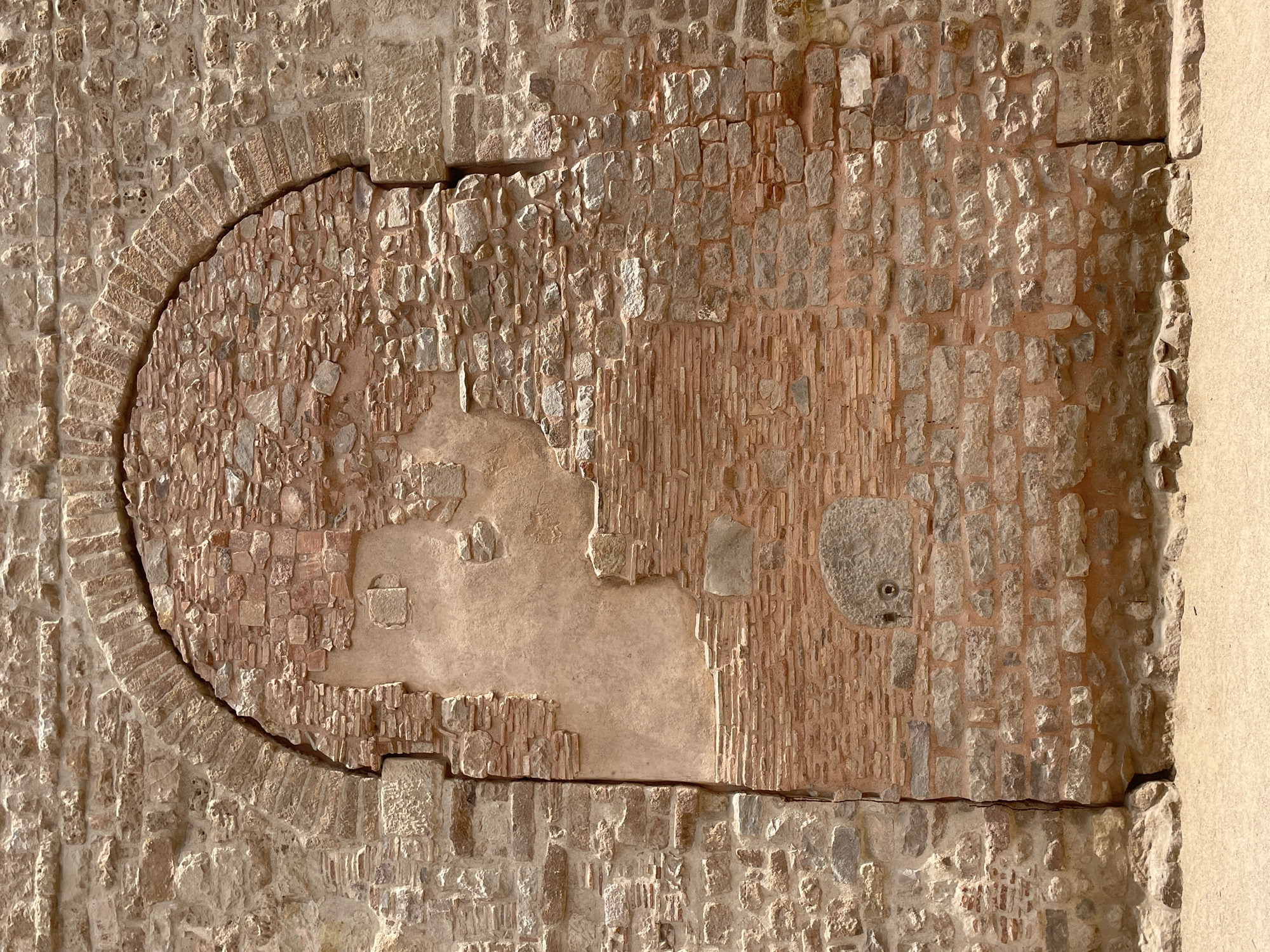 The image shows a weathered, arched stone entryway in Barcelona, Spain. The stone structure is a section of an old wall, with a large, arched opening that has been partially filled in with smaller bricks and stucco. The original stones are rough-hewn and vary in color from light tan to reddish-brown. Some areas show significant erosion and damage, with loose stones and crumbling mortar. The brickwork filling the arch is more uniform in color and texture, but also appears aged. A section of rough plaster is visible within the arch. The ground in front of the wall is covered in sandy dirt, and the lighting suggests it is daytime. There are no people or other objects visible in the frame.