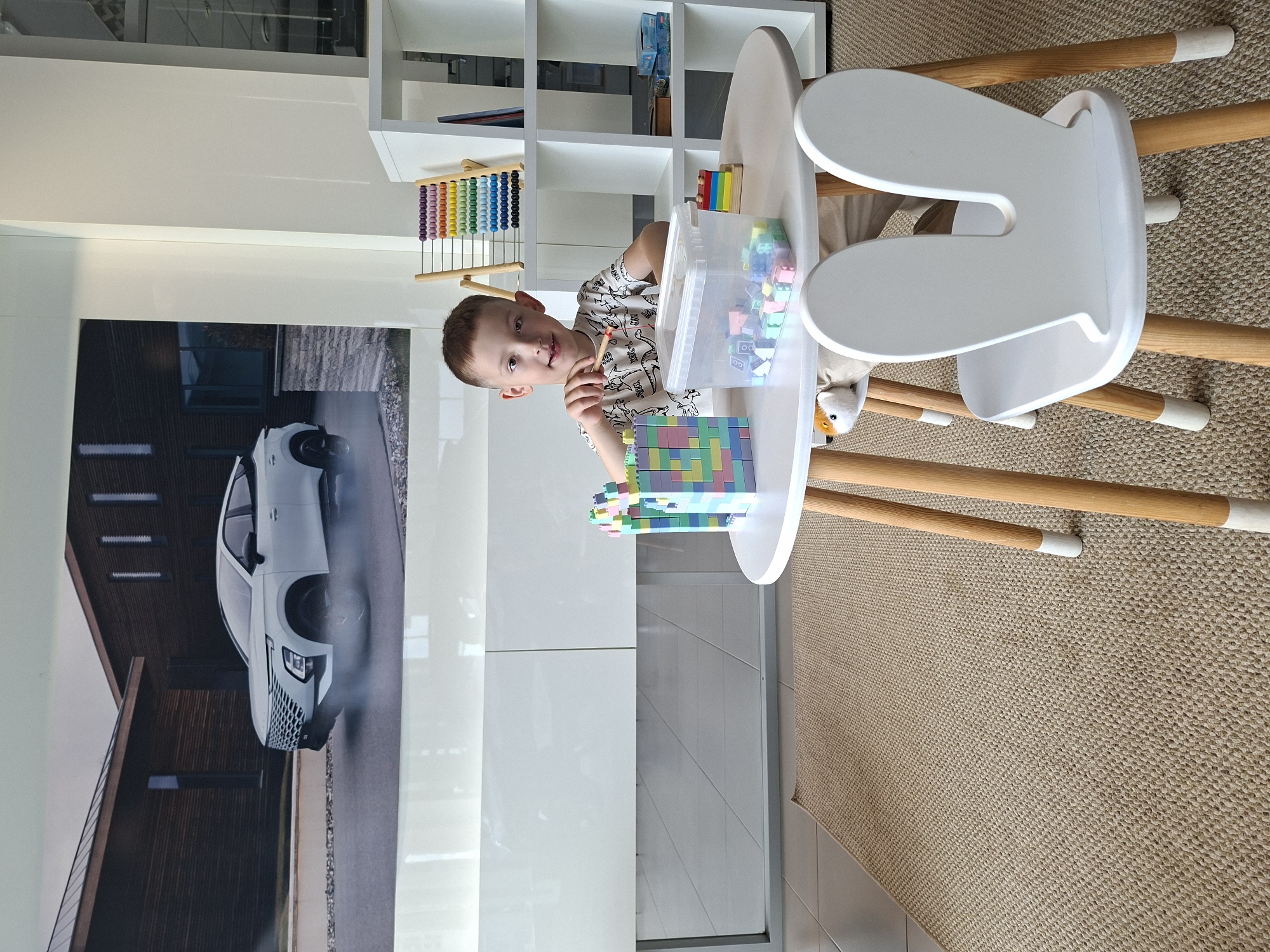 A young boy is playing with colorful building blocks at a white round table in a room that appears to be a showroom or play area. The boy is wearing a white t-shirt with a pattern on it and is looking directly at the camera with a slight smile. He is holding a small stick or pencil. In front of him on the table are building blocks, a clear plastic container filled with more blocks, and a partially built structure made of blocks. A white chair with a design resembling bunny ears is pulled up to the table. In the background, a large promotional image of a white car with dark accents is displayed on a wall, with wooden-paneled buildings behind it. A white shelving unit with an abacus and other items is also visible to the right. The floor is covered with a beige woven rug. The environment suggests a clean, modern space, possibly a car dealership's child-friendly waiting area, in the city of Izhevsk, Russia.