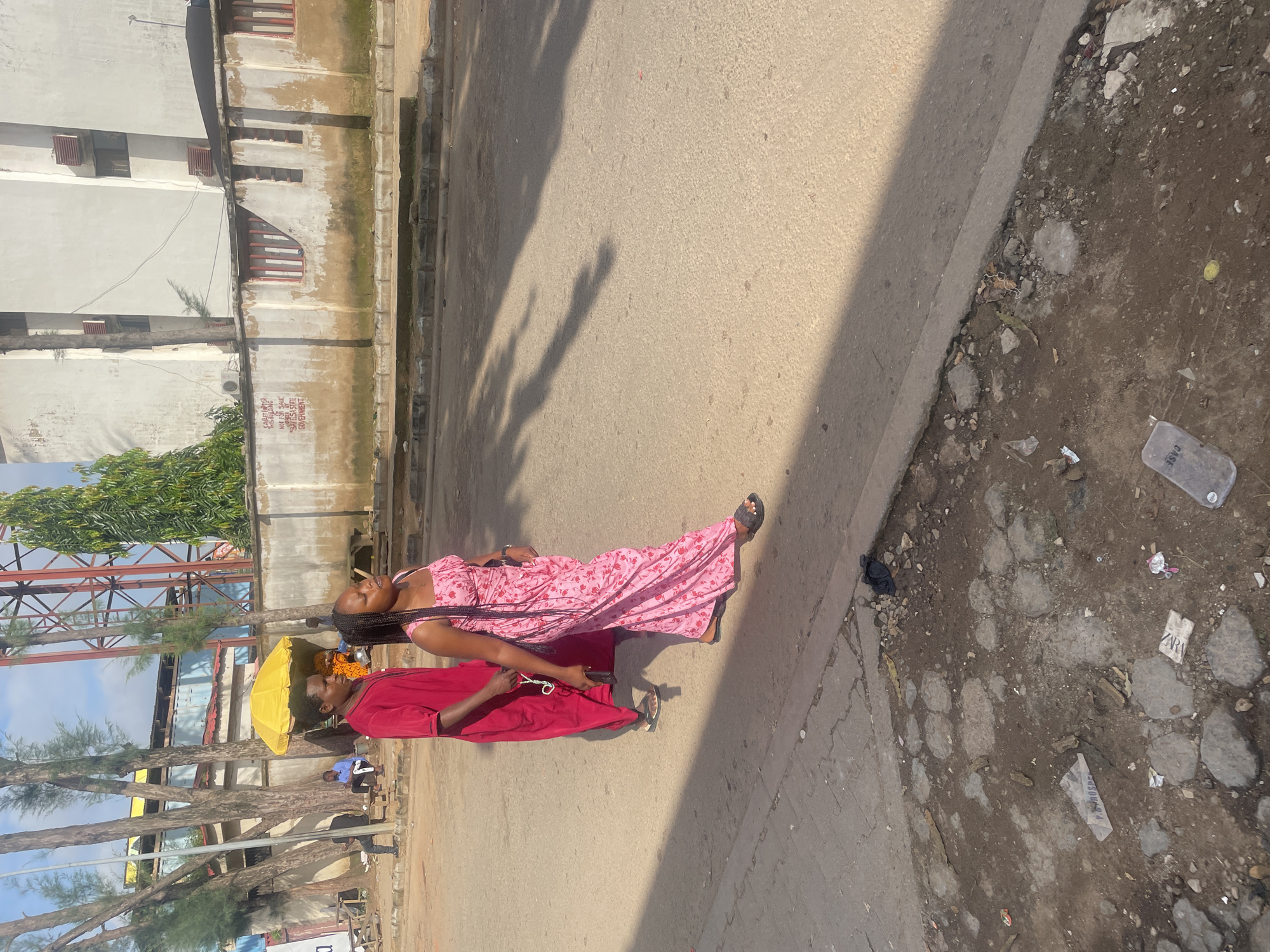Two individuals are walking on a paved street, likely in Abuja, Nigeria. The person on the left is wearing a red garment and appears to be looking forward. The person on the right is wearing a pink floral dress and looking to the side. They are walking away from the viewer towards a building with a white facade. In the background, there are trees, a yellow umbrella stand, and a metal tower. The street is lined with a concrete curb, and the ground beside the curb is rough and uneven with scattered debris. Sunlight casts long shadows across the street, indicating it is either morning or late afternoon. The scene appears to be an urban street, possibly near a marketplace or a residential area.