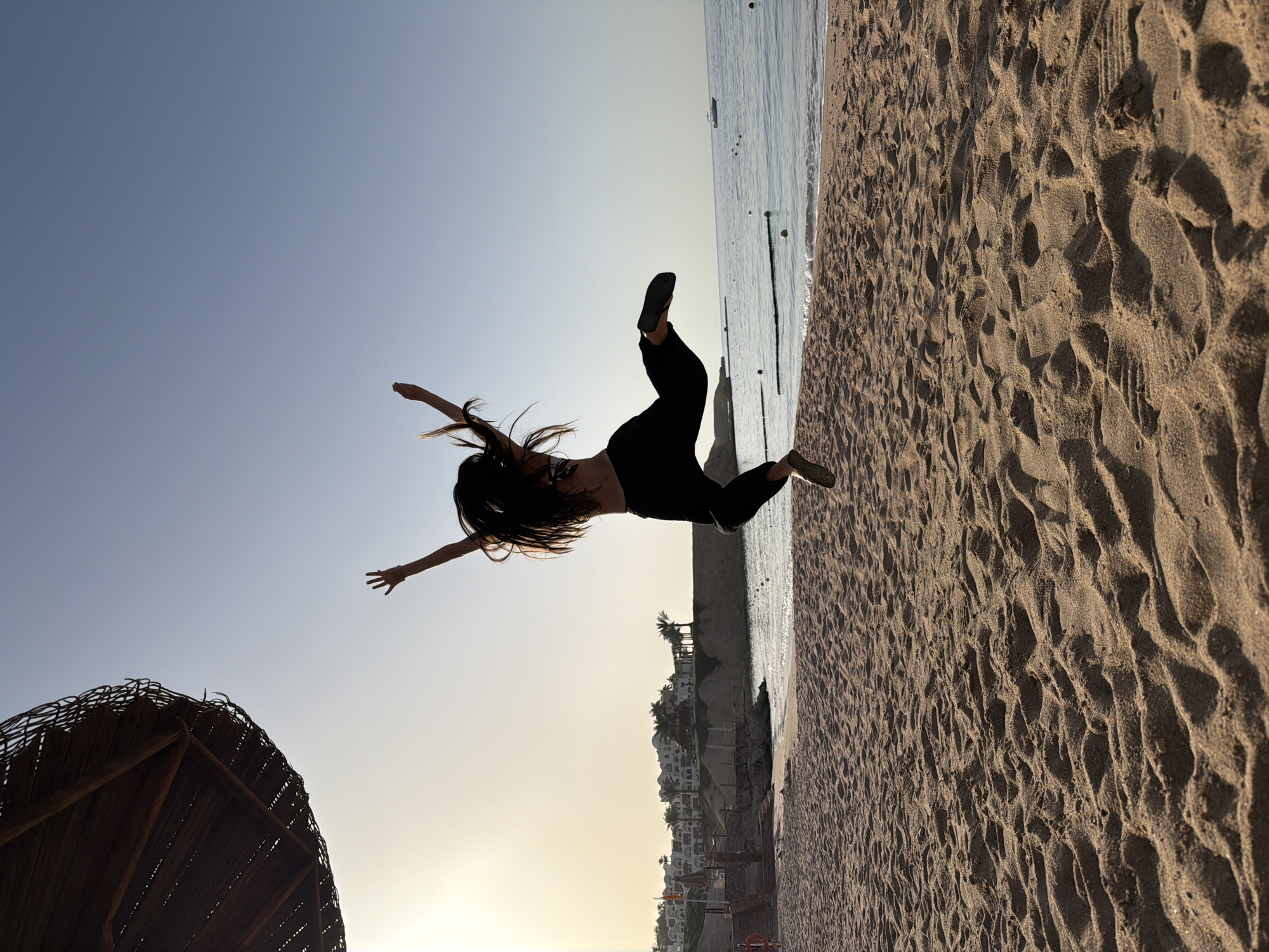 The image shows a woman leaping in the air on a beach at Sharm El Sheikh, Egypt. She has long dark hair, wears black pants, a black top, and shoes. Her arms are outstretched to either side. The sand is visible beneath her with the sea and buildings in the background. A beach umbrella is visible on the left side of the image. The sky transitions from blue to yellow.