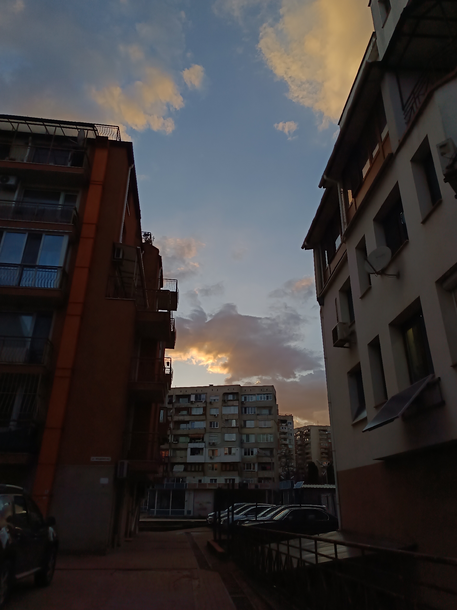 The image shows a narrow street view in Sofia, Bulgaria, framed by apartment buildings on either side. The sky is visible between the buildings, featuring a mix of blue and orange hues indicative of either sunrise or sunset, with clouds partially illuminated by the sun. The buildings are a mix of architectural styles, one with reddish-brown facade and visible balconies, and the other with a lighter, possibly off-white or beige facade, also showing windows and what appears to be satellite dishes and air conditioning units. Cars are parked along the street. The scene appears somewhat deserted, with no people immediately visible. The lighting suggests it is either early morning or late evening.