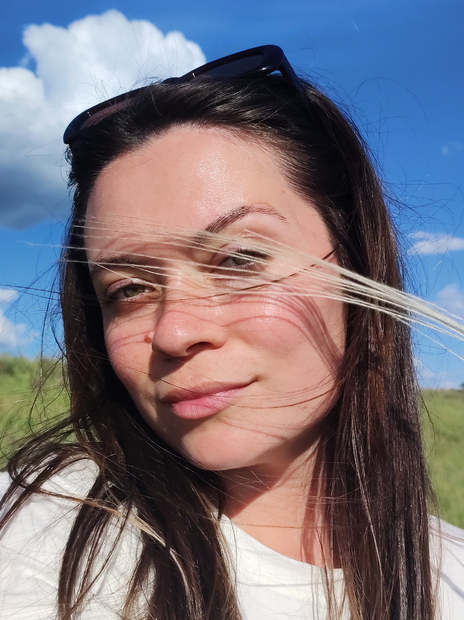 A close-up, natural-light portrait of a woman with brown hair and light skin, taken outdoors in Chaikovsky, Russia. The background features a blue sky with fluffy white clouds and some green vegetation. The woman is wearing sunglasses on top of her head and a white shirt. Her hair is slightly windblown. The photo appears to be taken during the daytime.