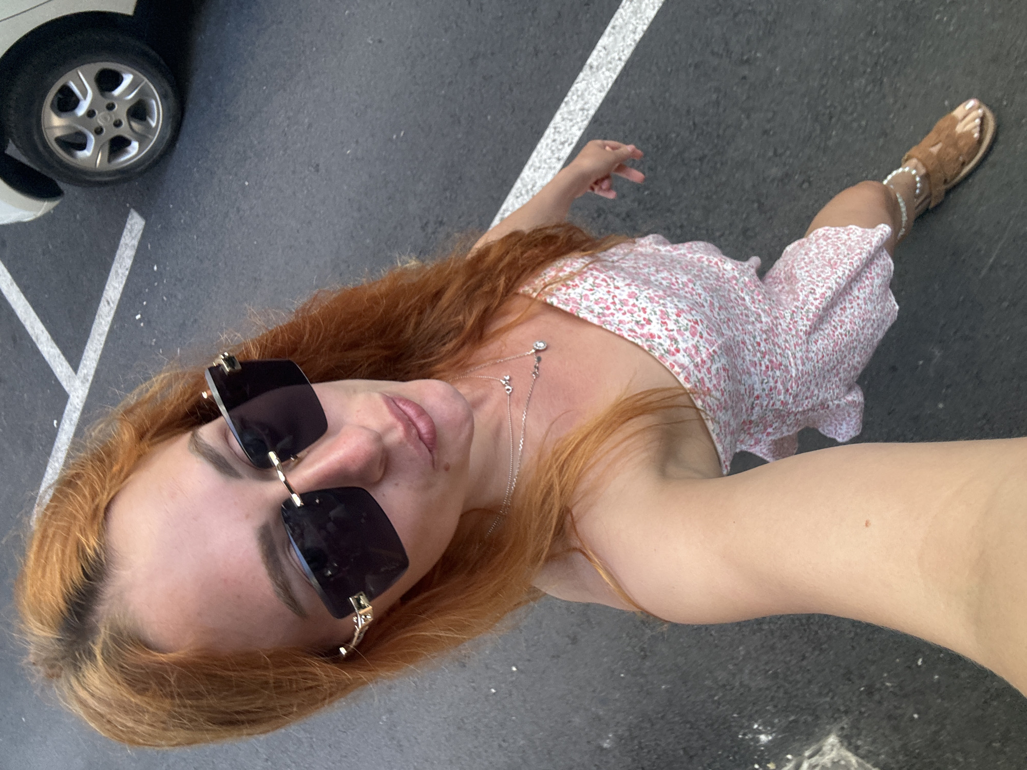 A high-angle selfie captures a female individual standing on a dark asphalt surface in Budva, Montenegro. The subject has long, reddish-orange hair with darker roots visible at the scalp. She wears large, dark, rectangular sunglasses with gold-colored metal accents at the temples, obscuring her eyes. Her lips are slightly parted in a soft pout. She is attired in a sleeveless, light-colored dress featuring a small pink floral pattern. Multiple delicate silver-colored necklaces are visible, one featuring a "love" script pendant and another with a small circular charm. A white beaded or pearl-like anklet is present on her right ankle, above a brown, multi-strap flat sandal. The individual's right arm is partially visible in the lower-left, holding the camera. The background consists of dark asphalt pavement marked with white painted lines, specifically a chevron pattern in the upper-left and a straight line extending across the mid-right. A white vehicle, partially visible in the upper-right quadrant, displays a silver multi-spoke wheel rim.