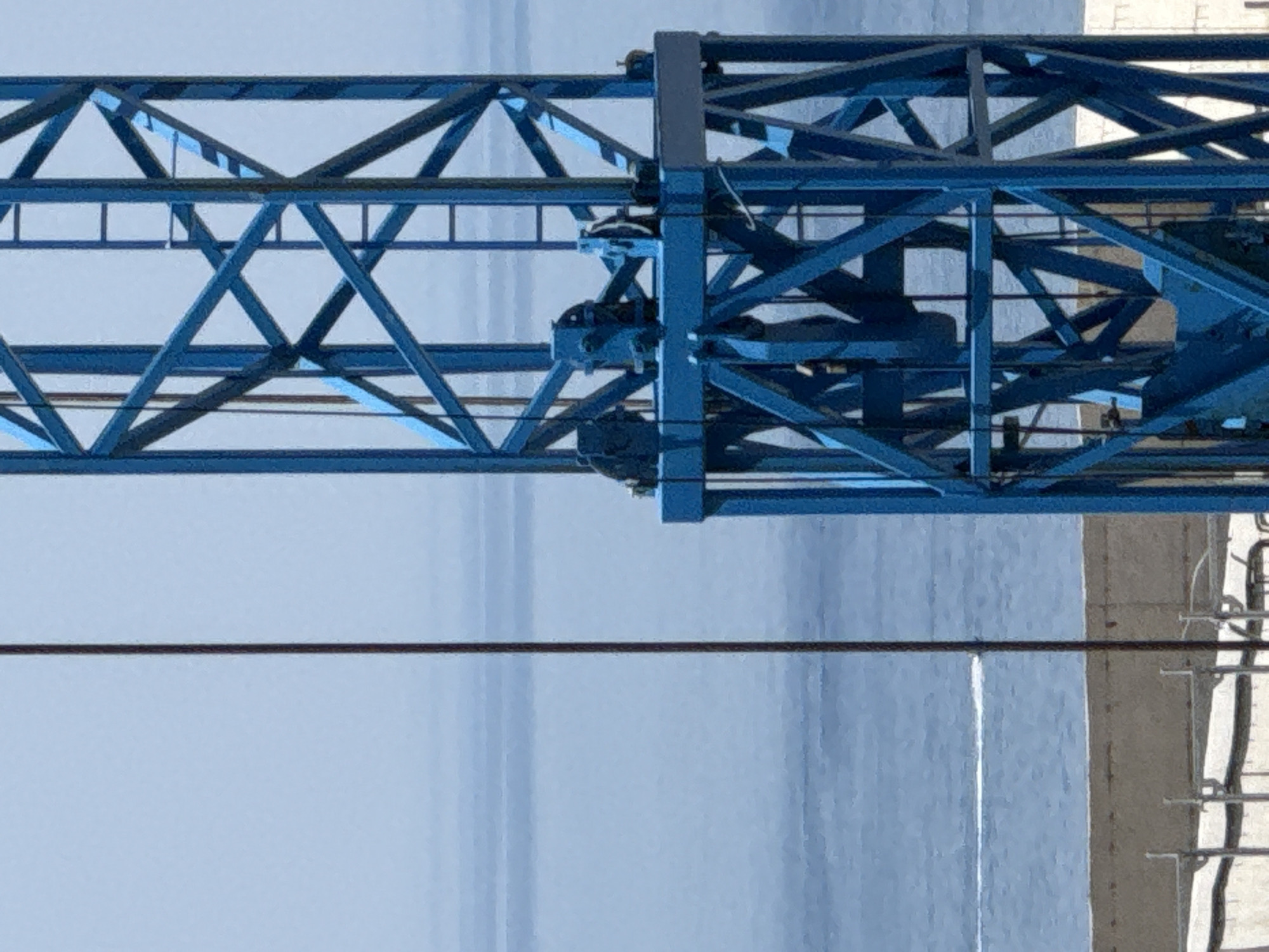 The image captures a close-up view of a blue construction crane against a hazy sky and a distant body of water, likely the Adriatic Sea. The crane's structure is made of intersecting metal beams, with visible pulleys and cables. A thin, dark cable runs vertically down the left side of the frame, obscuring part of the background. In the distance, a faint white trail on the water suggests the movement of a boat. The lower portion of the image shows a concrete structure and scaffolding, indicating ongoing construction. The scene is set in Bečići, Montenegro, a coastal resort town known for its beaches and hotels. No people are visible in the image.