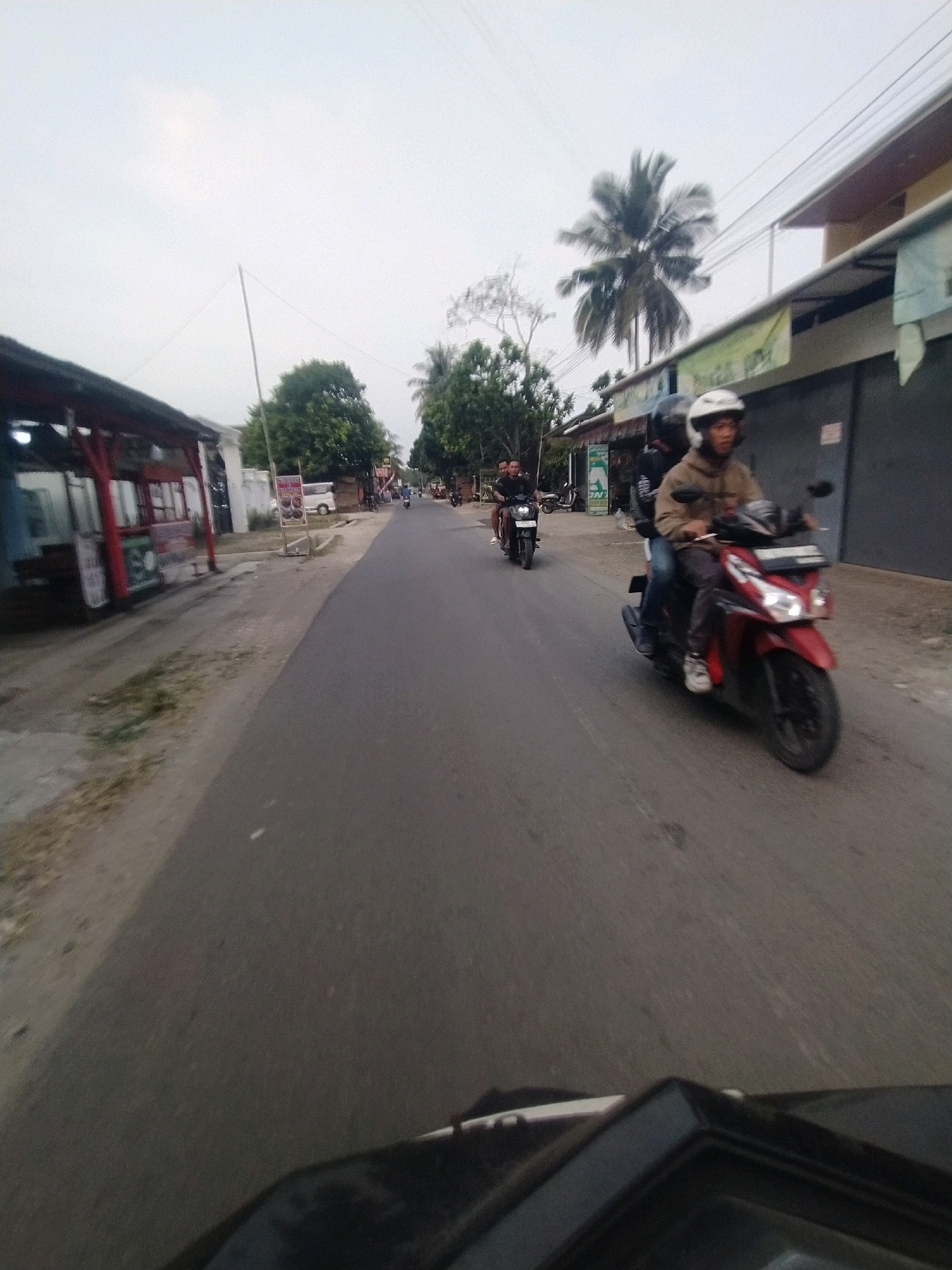 A street scene in Desa Karangjaladri, Indonesia, shows two motorcyclists on a road. The foreground is dominated by the top of a dark vehicle, likely a motorcycle, from which the photo is taken. On the right side of the road, a red scooter is ridden by a person wearing a helmet and a light brown jacket. Behind them, another person wearing a helmet is visible. The road is asphalt and appears to be in a semi-urban or rural area, with buildings and vegetation lining the sides. On the left, a covered stall with red posts is visible. Trees, including a palm tree, are scattered throughout the scene. The sky is overcast.