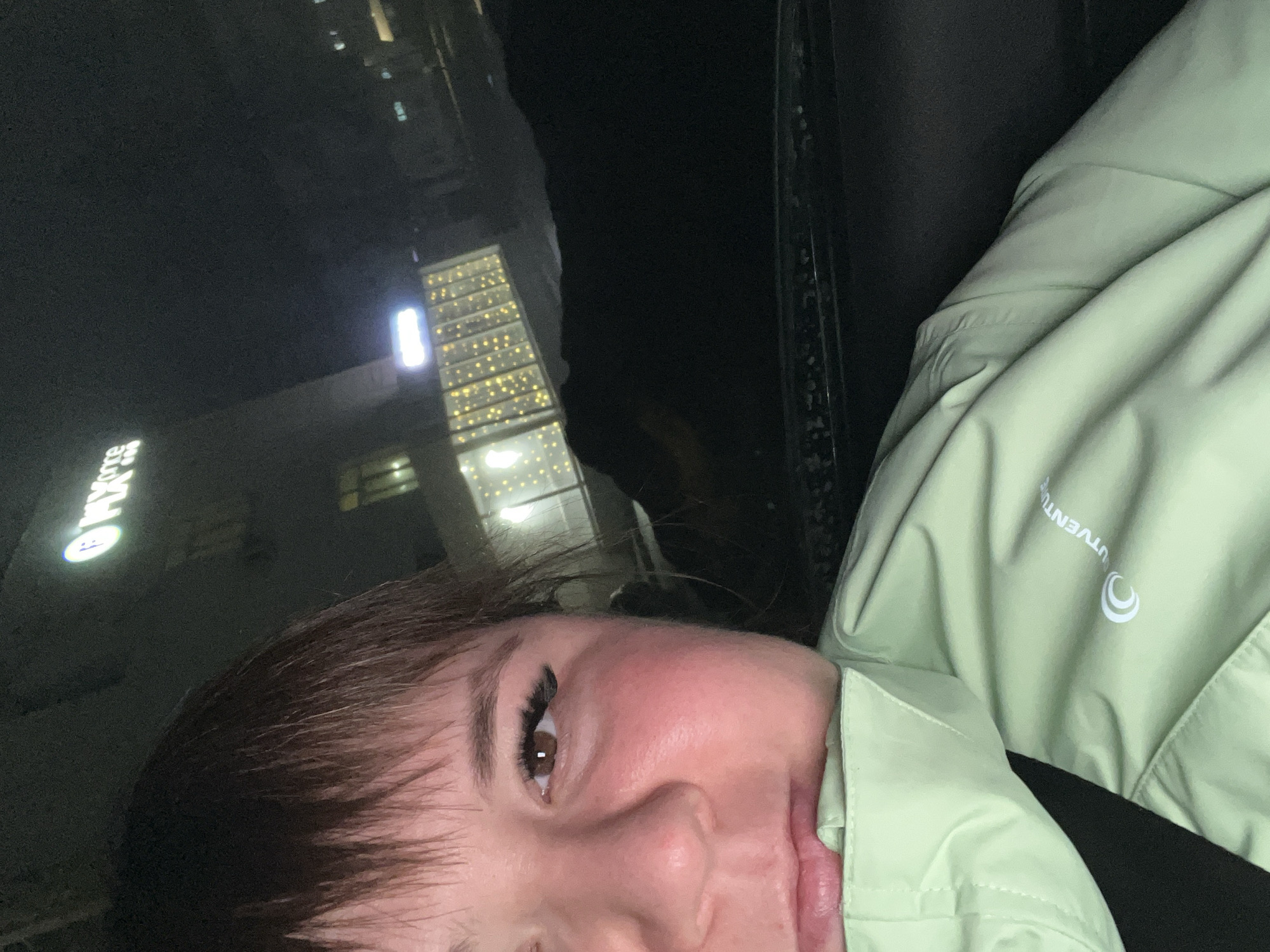 A close-up, low-angle shot shows a person's face, predominantly their eye, nose, and mouth. The person has brown hair with bangs and is wearing artificial eyelashes. Their skin is fair with a reddish tint on the cheek. The person is wearing a light green jacket with the word "UTVENTURE" visible on the right side.
In the background, through what appears to be a window, is a night scene of the city of Chaikovsky, Russia. Several buildings are visible, including one with illuminated windows and another with bright signs. The overall lighting is dim, typical of a night scene in a city.