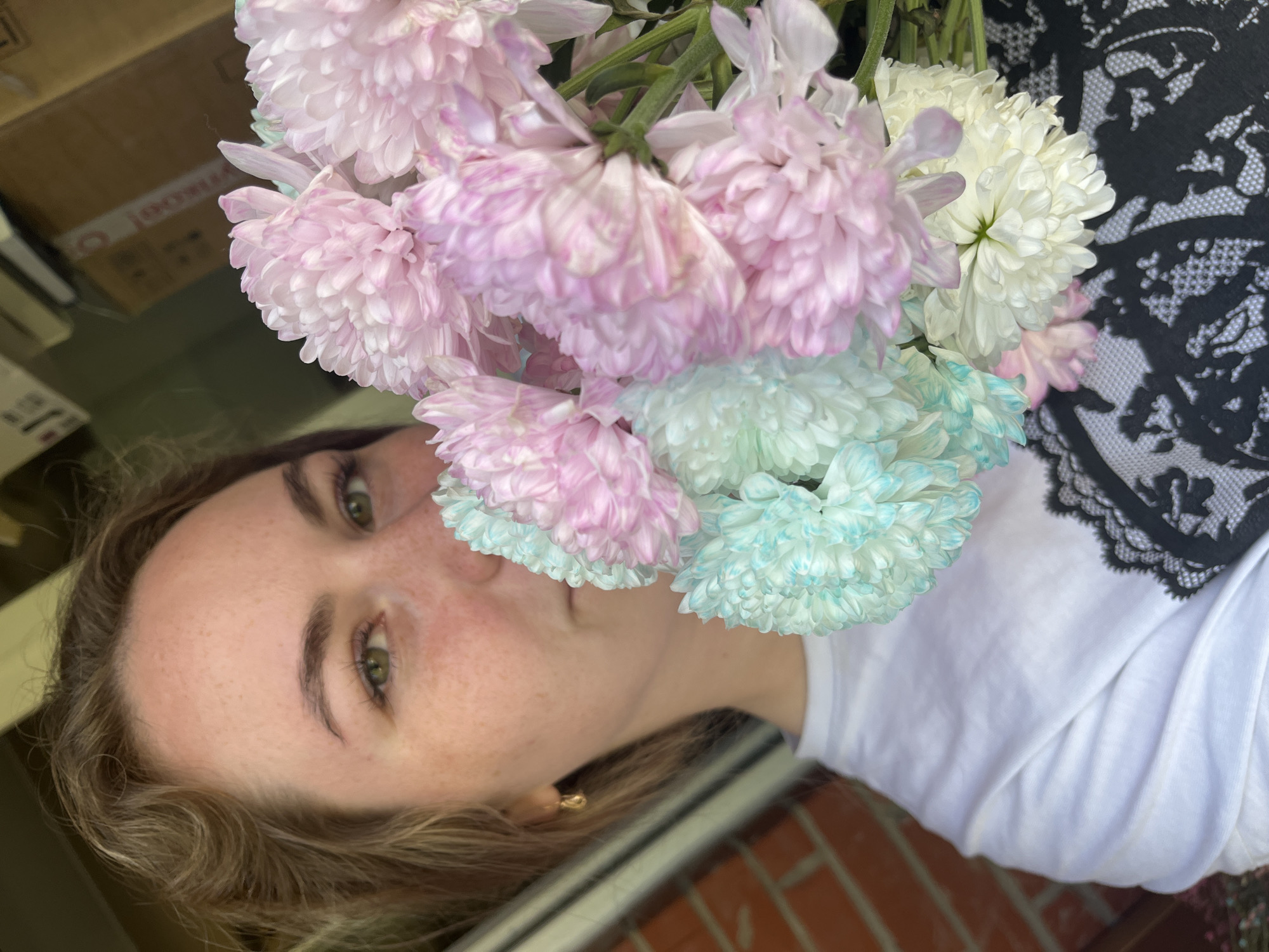 A young, fair-skinned woman with light brown hair and freckles is holding a bouquet of chrysanthemums. The flowers are a mix of pink, light blue, and white. She is wearing a white t-shirt and has a gold earring visible in her left ear. In the background, there is a cardboard box, a black lace garment, and a brick wall. The location cannot be identified as Chaikovsky, Russia based on the image.