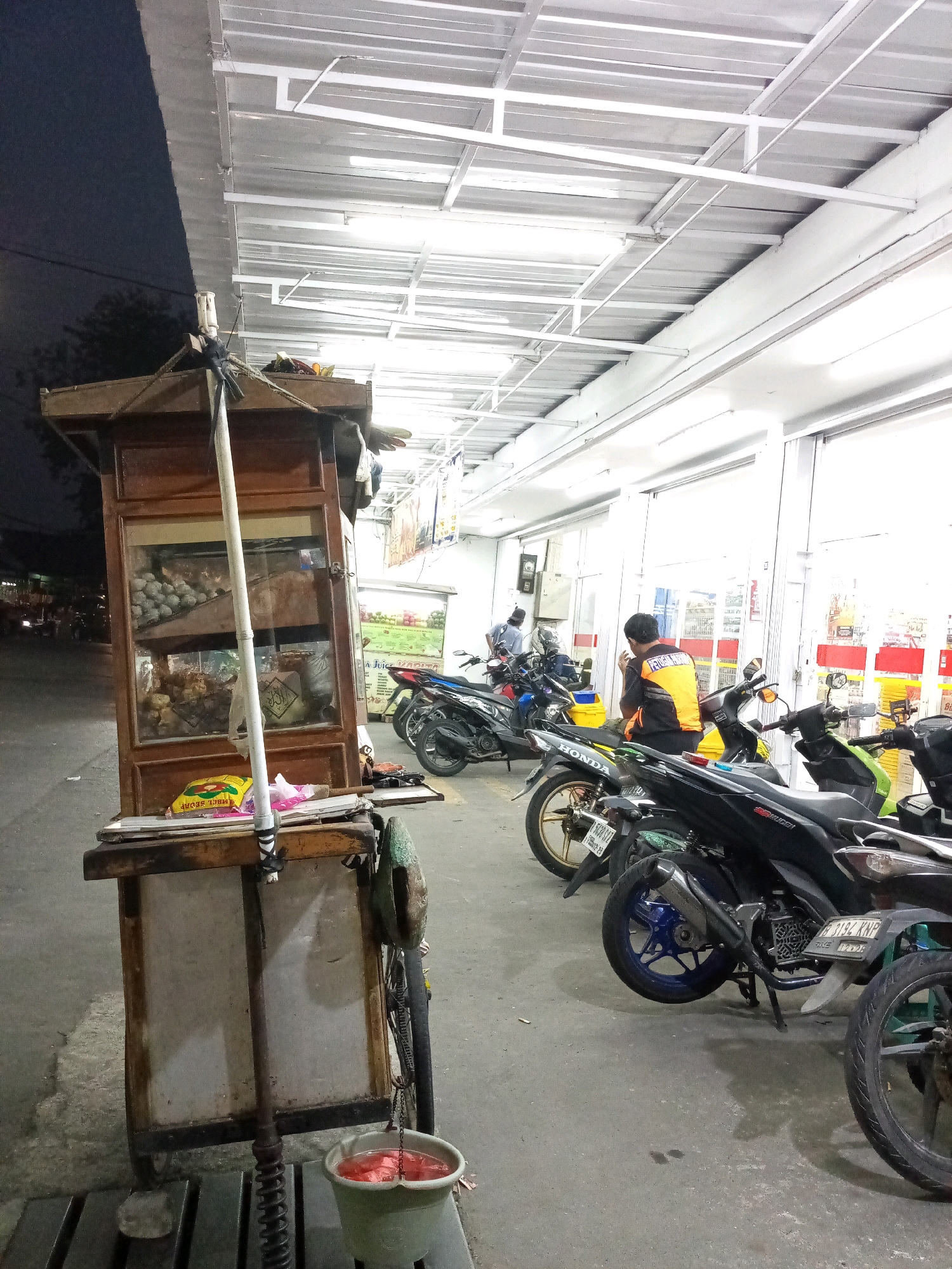 The image depicts a street scene in Bekasi, Indonesia, at what appears to be dusk or night. In the foreground, a wooden food cart with a glass display case is positioned on a concrete sidewalk. The cart contains various food items, including what look like balls of food in the upper section and other items in the lower section. A metal pole extends vertically from the cart, and a green bucket filled with a red liquid is hanging from a chain attached to the cart's base. Several motorcycles are parked in a row behind the food cart, some with their engines off and others potentially running.
Further back, a building with a white, corrugated metal roof and numerous fluorescent lights extends along the street. The building's facade appears to be made of glass panels, suggesting it might be a shop or convenience store. A person wearing an orange vest over a black shirt is standing near the entrance of this building, interacting with one of the motorcycles. Another person is partially visible inside the building. The street in front of the cart and motorcycles is paved with asphalt, and there are some discarded items on the ground, including a yellow packet labeled "MBL SEDAP". The overall environment suggests a typical urban setting in Indonesia, likely a commercial or residential area with street vendors and shops.
