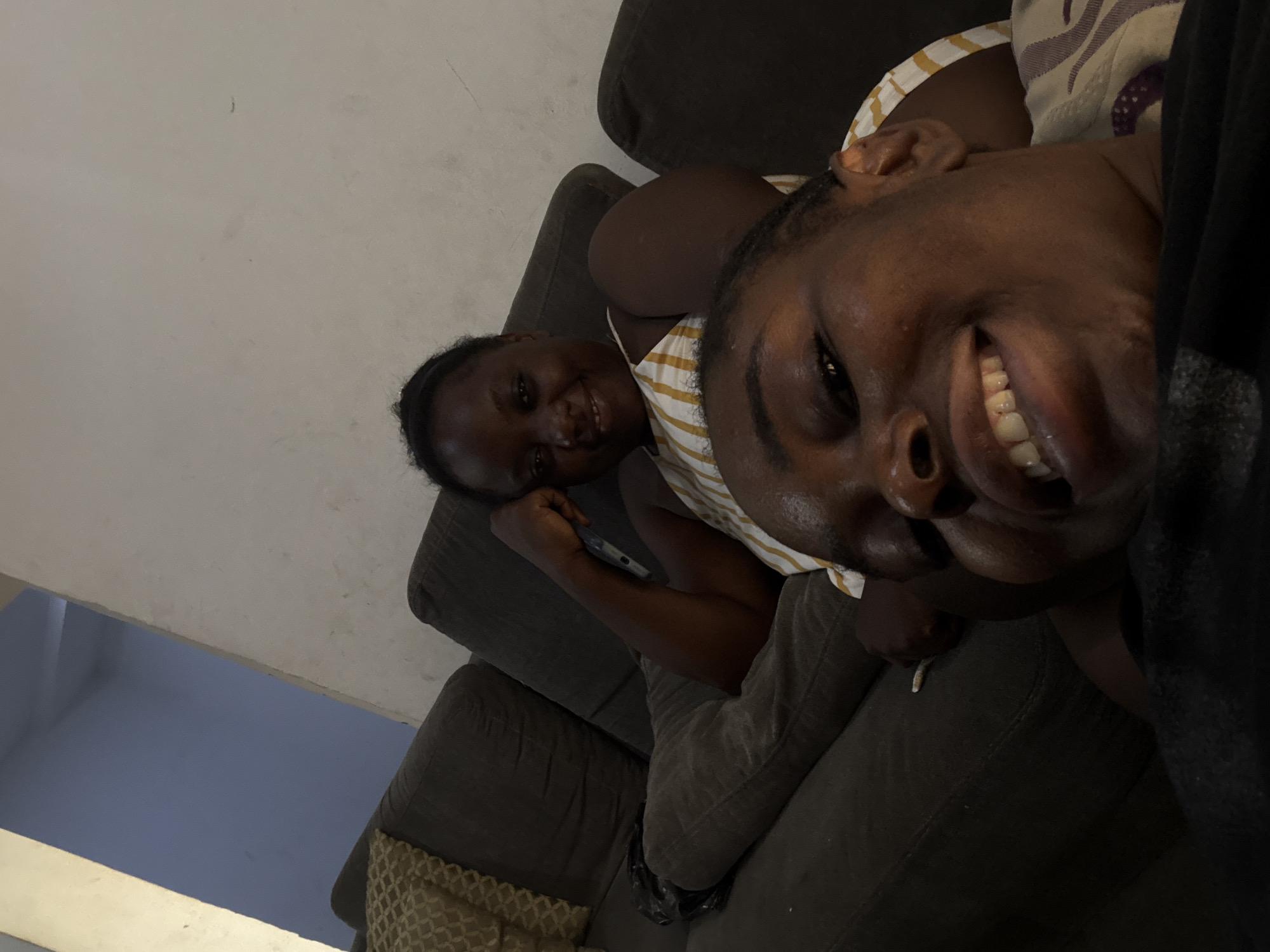 This intimate indoor photograph captures two individuals, likely young women, relaxing on a dark grey or brown sofa. The camera perspective is angled from above, creating a close-up, selfie-like shot.
In the foreground, largely occupying the right side of the frame, is one person with dark skin, smiling broadly at the camera, revealing white teeth. Only their right eye, nose, mouth, and parts of their cheek and forehead are visible. They appear to be wearing a dark, possibly textured, top.
Further back and to the left, the second person, also with dark skin, is fully visible from the waist up, lying on the sofa. They are wearing a striped top, likely yellow and white, and are also smiling cheerfully towards the camera. Their left hand holds a dark-colored mobile phone near their chest.
The sofa is upholstered in a dark fabric. A patterned, possibly gold and dark, cushion or throw is visible on the sofa in the lower left. The upper left portion of the image shows a plain, light-colored wall. A darker blue area is visible in the bottom left corner, possibly a lower part of the wall or a detail of the room. Both individuals express happiness and comfort, suggested by their relaxed postures and wide smiles. The setting suggests a home environment in Abuja, Nigeria, and the lighting indicates an indoor shot, possibly in the evening. There is no visible text.
