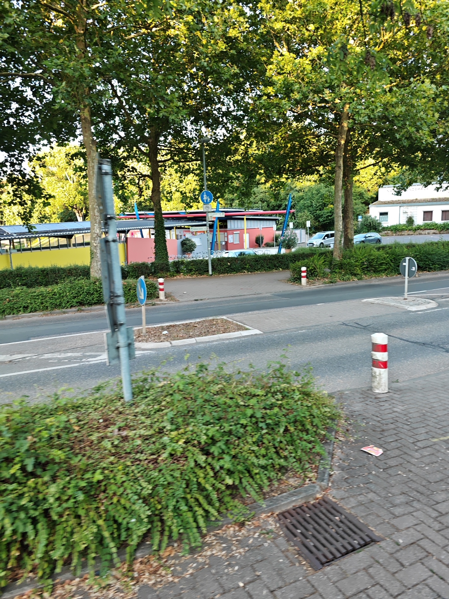This is an exterior, eye-level, full-shot photograph taken in Nordheim, Germany. The image captures a scene along a road with a building and trees in the background. In the foreground, a paved sidewalk with a metal grate drain is visible, with a dense green bush bordering it. A bollard with red and white stripes stands on the sidewalk. The road surface is visible, with lane markings and a pedestrian crossing indicated by a raised curb and patterned paving stones. Further back, a low hedge lines the road, separating it from a pedestrian walkway. A light pole with traffic signs, including a circular blue sign with a bicycle and pedestrian symbol, is positioned near the walkway. A building with a yellow and red facade is partially visible behind the hedge, with a white building with windows and a pitched roof visible further in the background. Several trees with dense green foliage are prominent throughout the scene, with sunlight filtering through their leaves. Two cars are parked on the road in the mid-ground. The overall scene suggests a quiet, possibly residential or commercial area, during daytime.