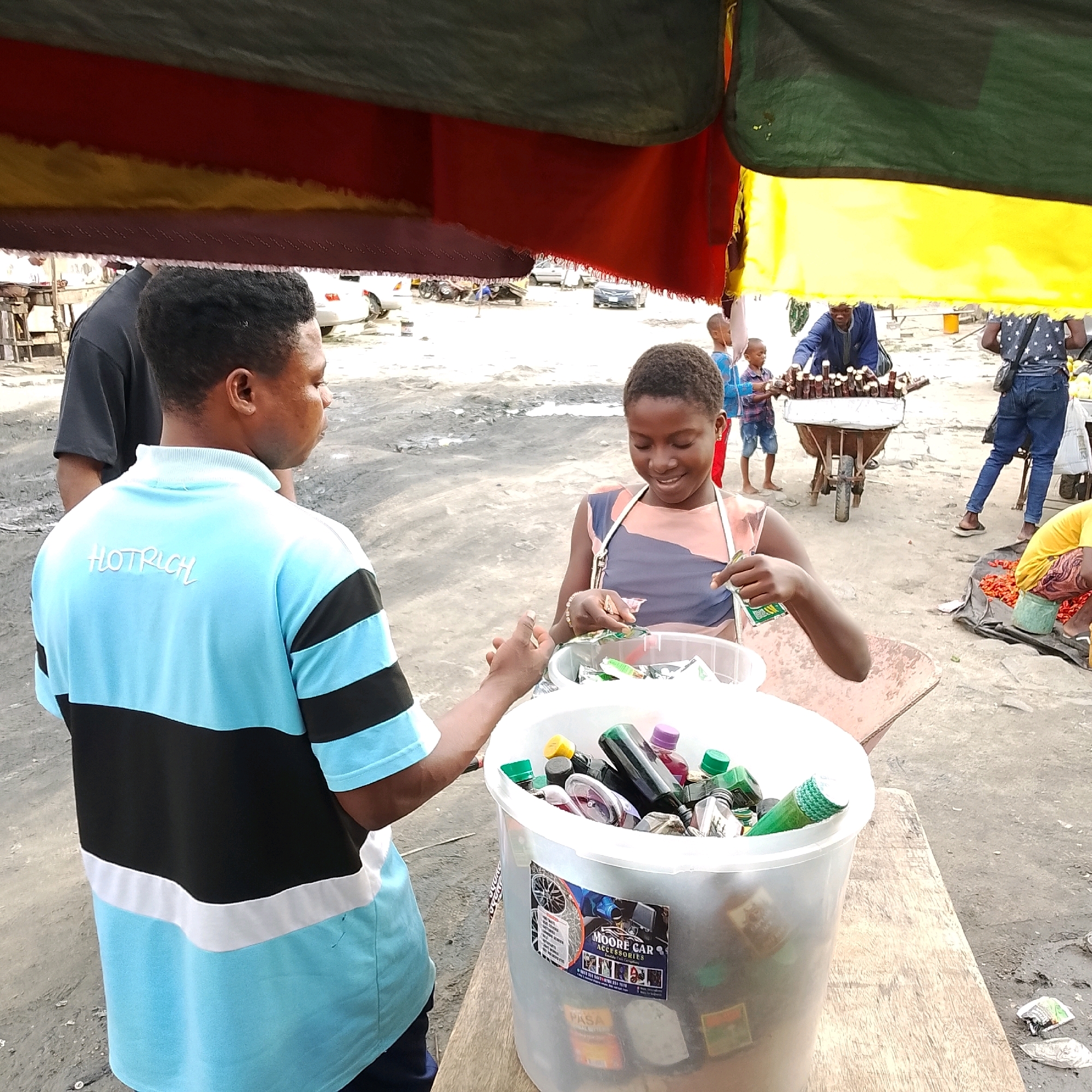 The image depicts an outdoor market scene in Ado, Nigeria, under a colorful awning of red, yellow, and green. In the foreground, a young person wearing a light blue and black striped polo shirt with "HOTRICH" visible on the back, stands next to a large white bucket filled with various small bottles of liquids. The person is interacting with a young girl who is smiling and holding a small packet. The girl is wearing a sleeveless, multi-colored dress and has a white strap across her shoulder. The bucket has stickers on its side, one prominently displaying "MOORE CAR ACCESSORIES".
In the background, a man in a blue jacket is arranging rows of small brown bottles on a cart. Several children are visible in the middle ground, including a young boy in a blue shirt and denim shorts standing near the cart. A person in a dark shirt stands to the left, and another person in a grey shirt with a star pattern and denim jeans is visible to the right, with a black bag slung over their shoulder. Further in the background, cars and motorcycles are parked on a dirt and gravel surface, typical of an urban Nigerian environment. A vendor in a yellow shirt and patterned pants is crouched on the ground to the right, attending to a pile of red peppers on a black tarp. The overall environment suggests a busy, informal marketplace.