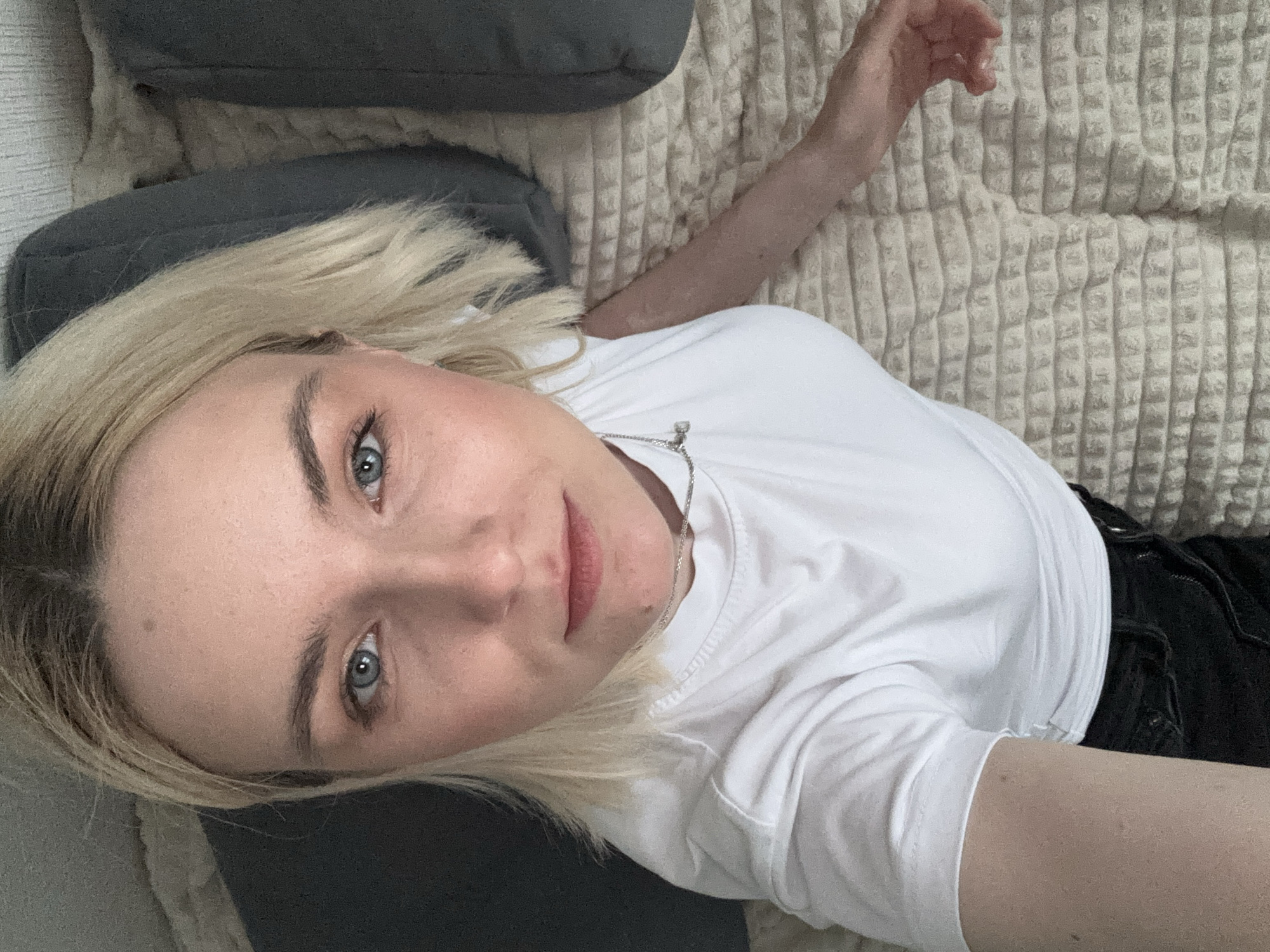 A young woman with blonde hair and blue eyes is lying on a beige textured blanket. She is wearing a white t-shirt and dark-colored pants or shorts. Two grey pillows are visible near her head. The image appears to be a selfie taken from above, looking down at the woman. There is no indication of location in Vsevolozhsk, Russia, or any other specific city.