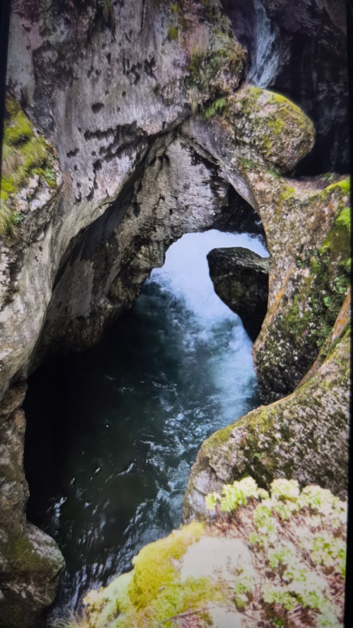 The image captures a dramatic aerial view looking down into a deep, narrow gorge or canyon, where a river flows through natural rock formations. The rugged rock walls, varying in shades of dark gray, brown, and reddish tones, are covered in patches of vibrant green moss and sparse vegetation, particularly along their upper edges.
The river water is a central feature, displaying dynamic movement. In the foreground and deeper sections, the water appears dark, almost black, with subtle ripples. Further into the gorge, where it passes through a natural archway formed by the rock, the water becomes a lighter, turbulent white and frothy blue, indicating a swift current or rapids. A prominent, moss-covered rock protrudes into the water from the right side of the arch, further constricting the flow.
The scene is lit in a way that suggests either an overcast sky or the depth of the gorge creating significant shadows, highlighting the illuminated, turbulent water as it emerges from the arch. No people, man-made objects, or visible text are present. The image conveys the powerful and ancient process of water carving through stone, showcasing the raw beauty of a natural landscape.