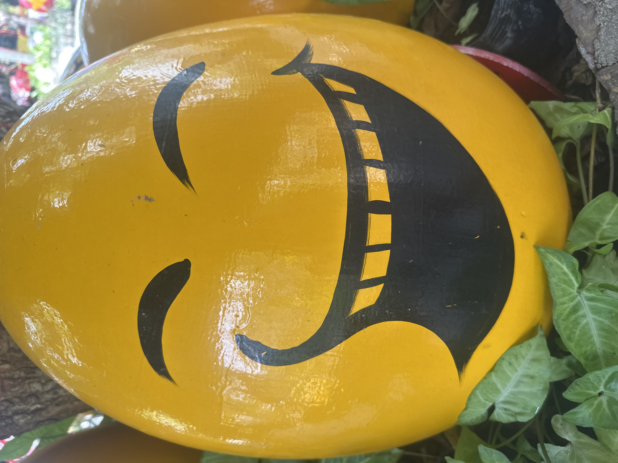 A close-up shows a round, yellow object resembling a large painted rock or decorative item. It has a black, cartoonish face painted on it, evoking a smiling or laughing emoji. The "mouth" is unusually large and features rectangular cutouts or "teeth." The object is situated outdoors, surrounded by greenery including small, heart-shaped leaves. The location is in Nam Nha Trang Ward, Vietnam.