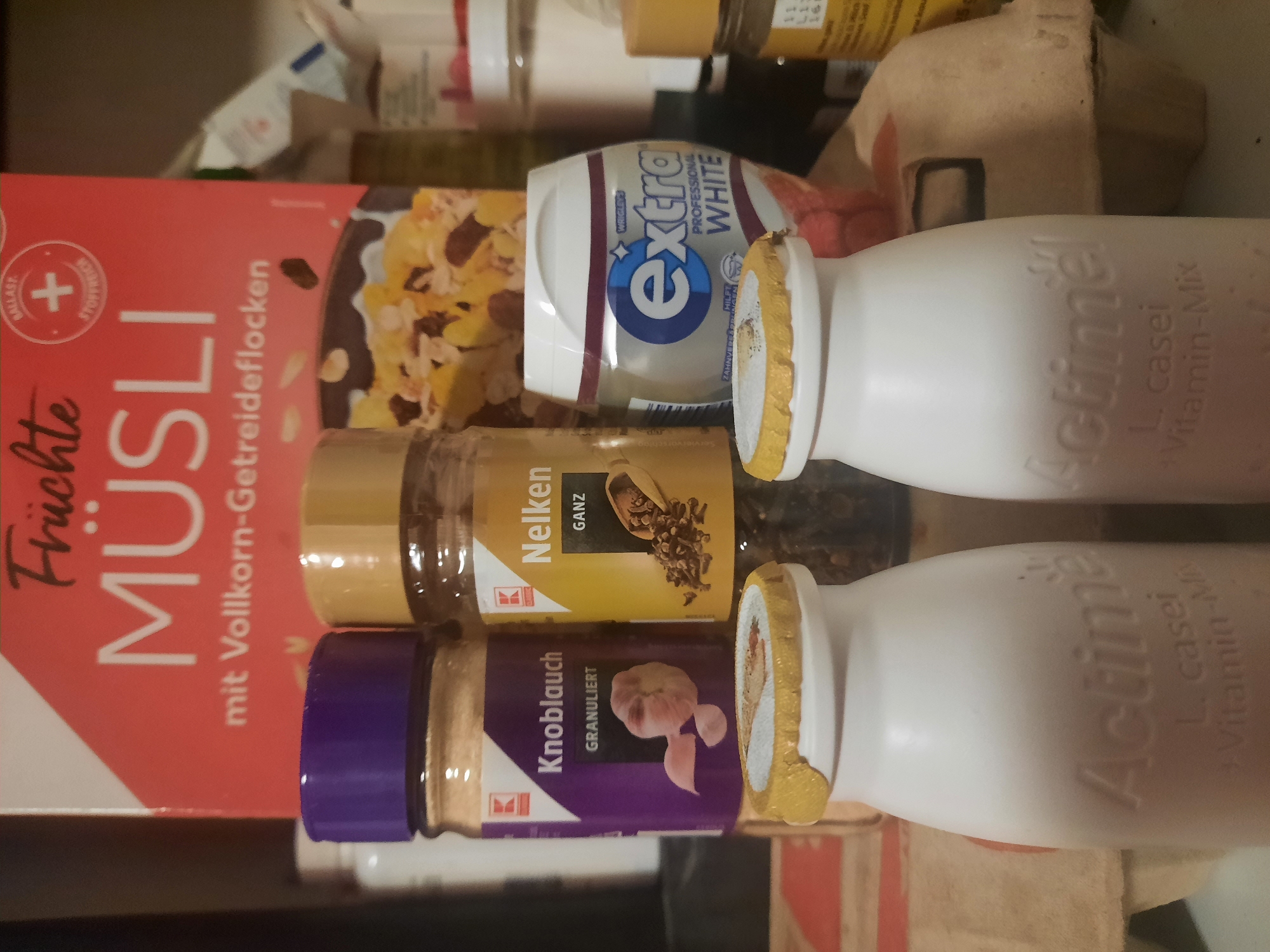 The image shows a collection of food and personal care items on a cluttered surface. In the background, a box of "Früchte Müsli" is visible, featuring text in German. In front of the muesli box, there are two spice jars: one labeled "Knoblauch GRANULIERT" with an image of garlic, and another labeled "Nelken GANZ" with whole cloves. To the right of the spice jars is a white and blue container of Wrigley's Extra Professional White chewing gum. In the foreground, two white bottles with gold caps are prominent, each bearing the embossed text "Actimel L. casei Vitamin-Mix." The overall setting appears to be a domestic environment, possibly a kitchen counter or shelf, with other items partially visible in the background. There is no identifiable information indicating a specific location in the city of Rosenheim, Germany. The lighting is dim, and the focus is primarily on the items in the foreground.