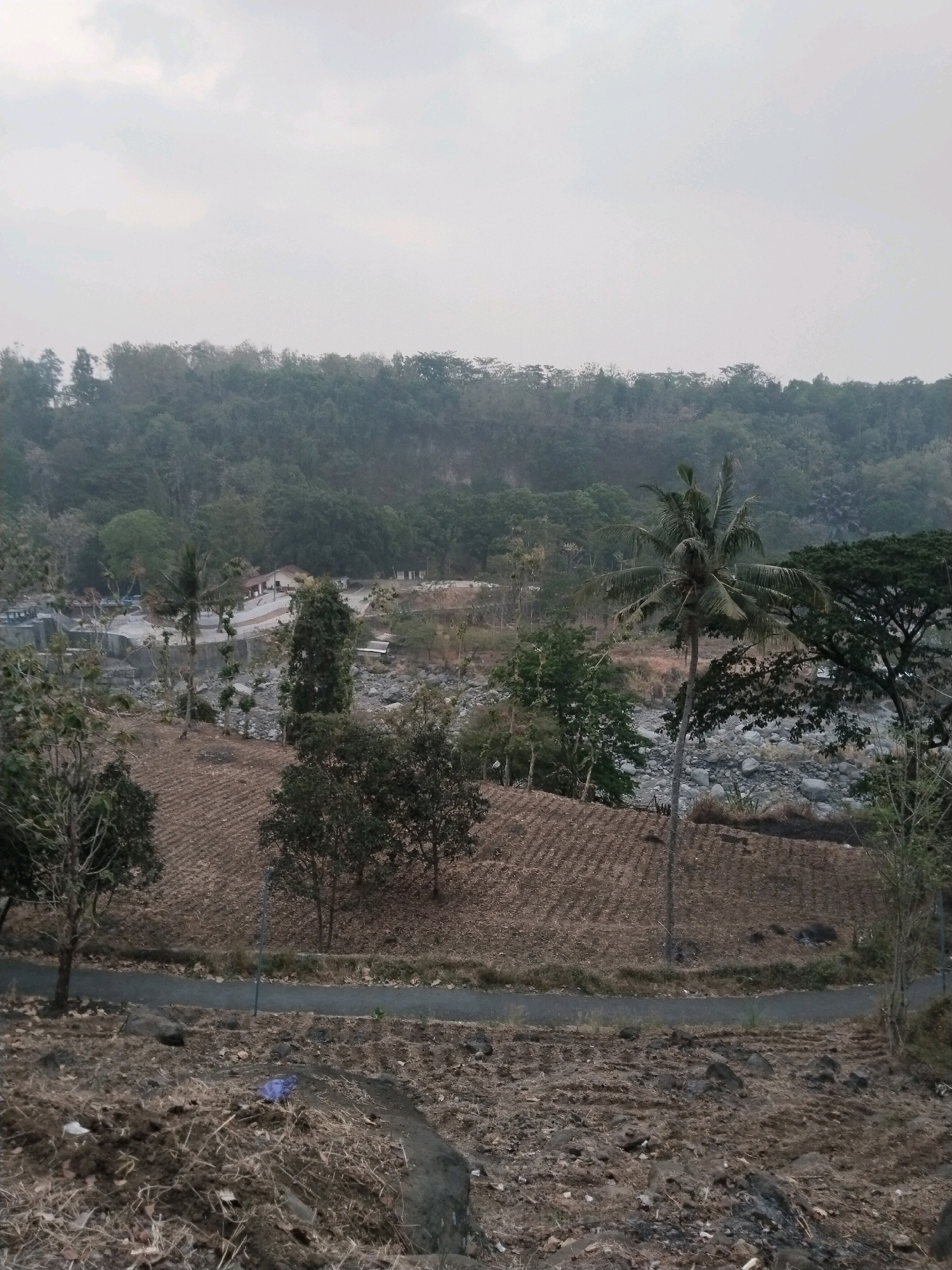 The image depicts a rural landscape in Sangkanjaya, Indonesia, characterized by a dry, rocky riverbed and terraced farmland. In the middle ground, a small cluster of buildings, possibly a resort or a housing complex, is visible next to the river. A road winds through the scene, connecting different areas. Lush green trees and a prominent palm tree punctuate the otherwise arid terrain. The sky is overcast, suggesting a cloudy day. The foreground shows dry soil with scattered rocks and vegetation. A blue plastic bag lies on the ground in the lower left portion of the image. The scene is tranquil and appears to be a natural setting with some human development.