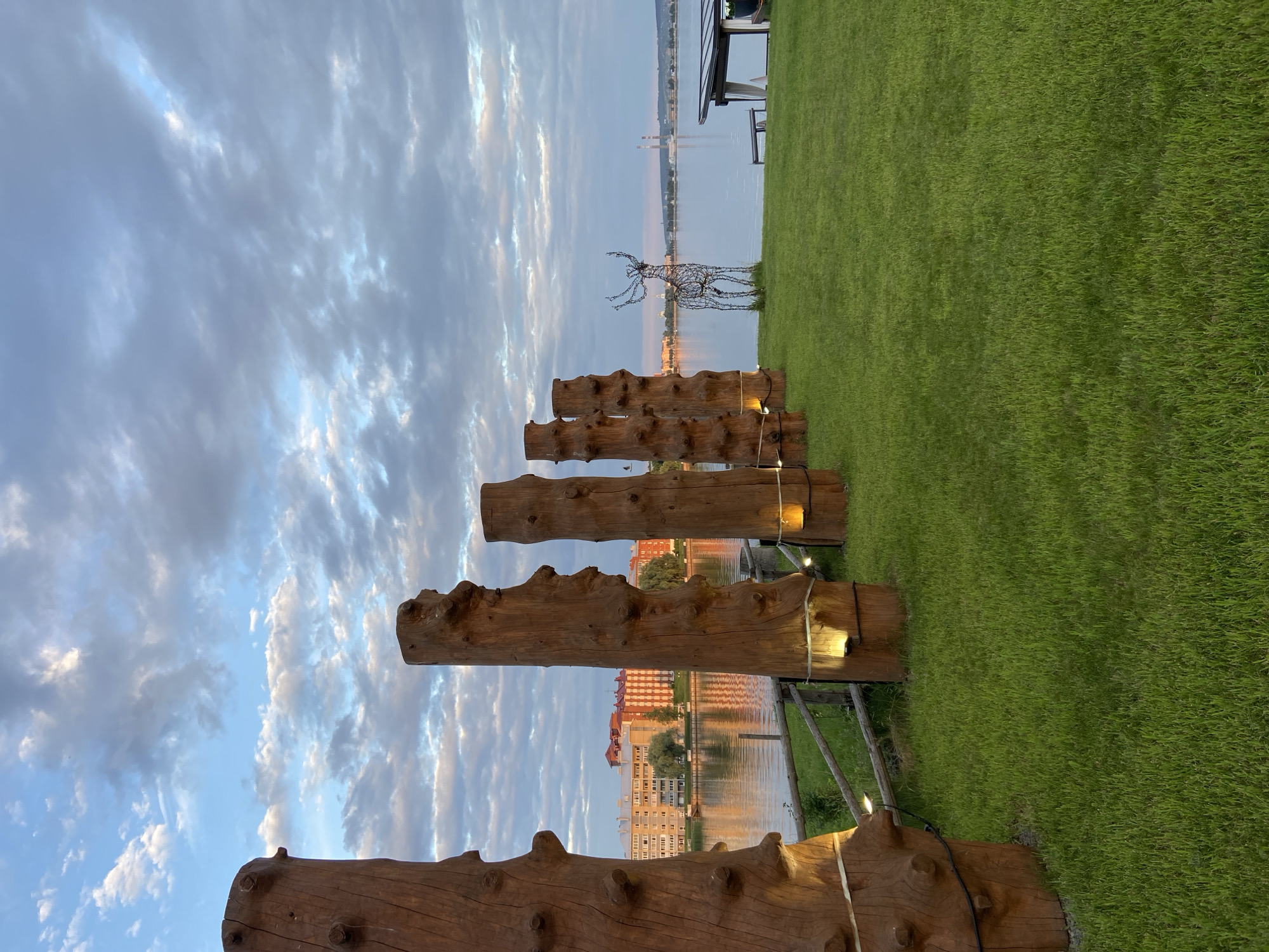 The image depicts a waterfront area in Votkinsk, Russia, likely near the Kama River, featuring a manicured lawn and several large, decorative wooden posts. These posts are illuminated from below with warm-toned lights and appear to be arranged in a line parallel to the water. In the background, a body of water reflects the sky and the lights of a city across the river. On the far bank, buildings with red and beige facades are visible. To the right of the wooden posts, a wire sculpture of a deer stands on the grass. Further in the distance, industrial chimneys and a small covered seating area with a white canopy are present. The sky is filled with a mix of light and dark clouds, suggesting either early morning or late evening. The overall scene conveys a sense of a well-maintained public or recreational space.