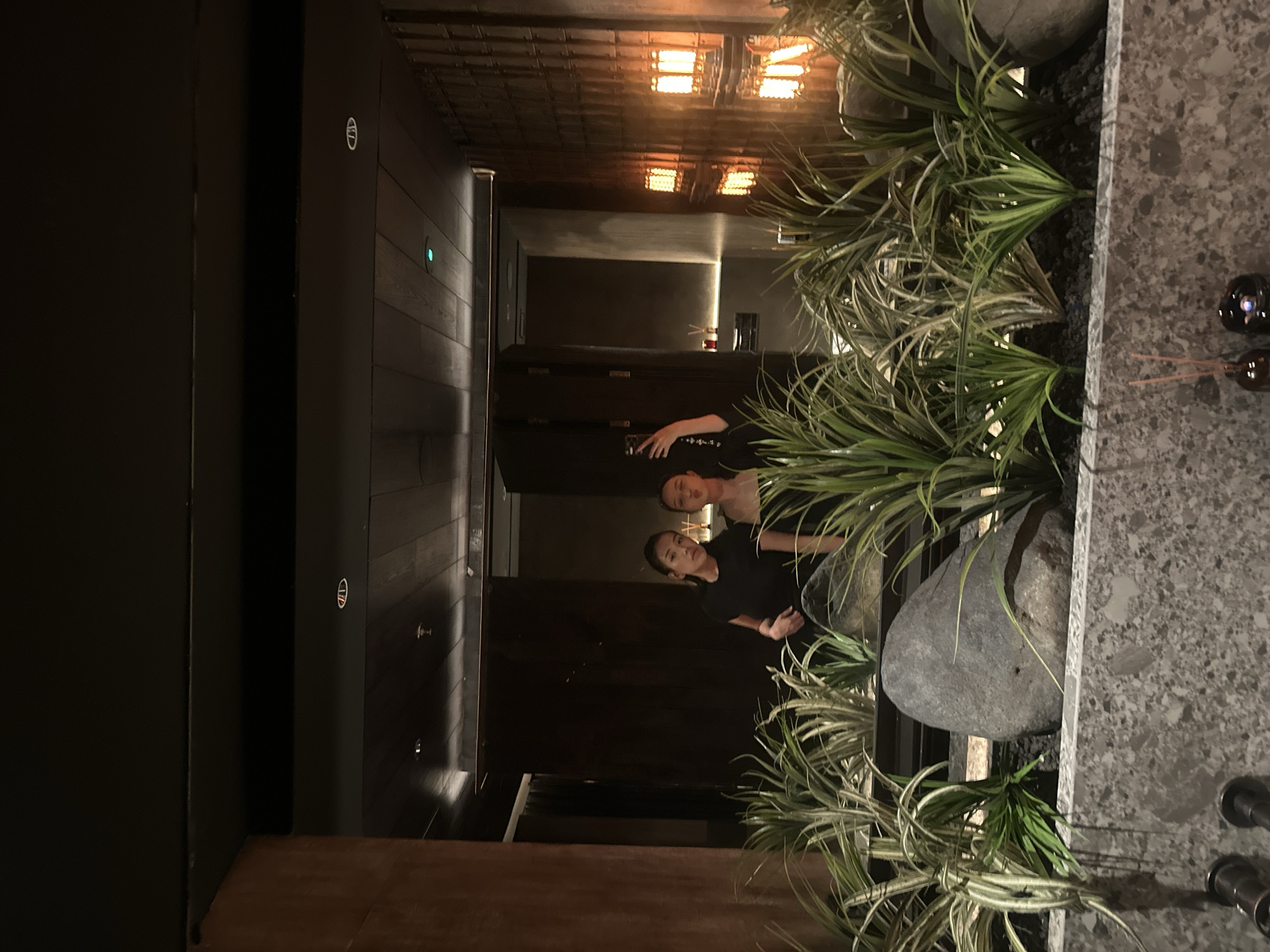 Two women are reflected in a surface that appears to be part of an indoor corridor or entrance area. The surface reflects a view down a dimly lit hallway with wooden paneling and ceiling lights. The women are standing to the side of the surface, and one is holding a cell phone. The area next to the reflecting surface includes decorative elements such as plants and a large stone. The image was taken in North Kuta, Indonesia.