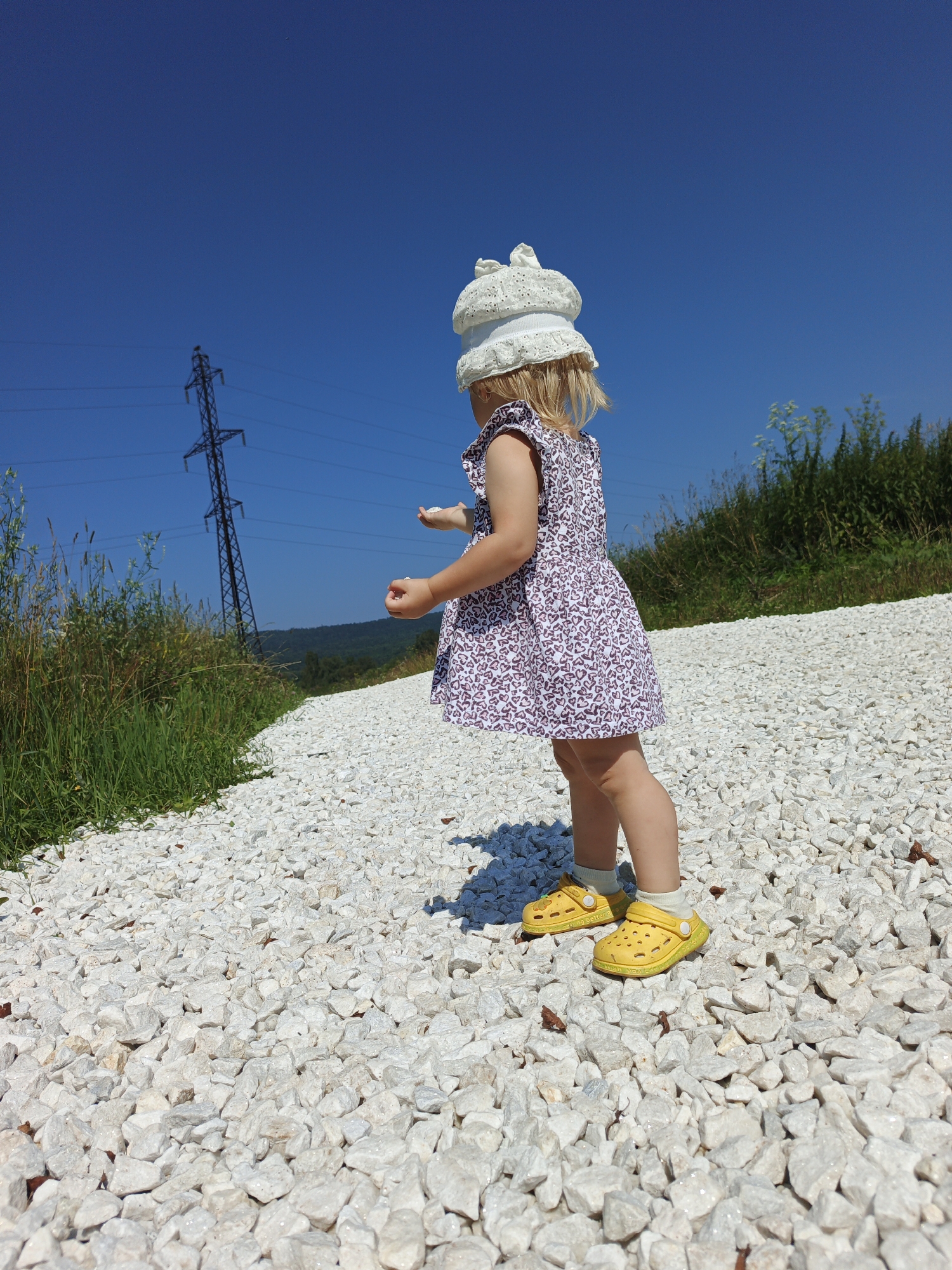 A young girl, viewed from the back and side, stands on a gravel path in a rural outdoor setting. She is wearing a white lace hat, a purple and white floral dress with small heart patterns, white socks, and yellow clogs. The girl is holding small white objects in her hands, possibly pebbles. The background features a bright blue sky, a metal power line tower, green grass, and distant hills. The path is covered in light-colored, irregularly shaped gravel. The image is taken from a low angle, emphasizing the child and the textured ground. The location is identified as Urtiushka, Russia.