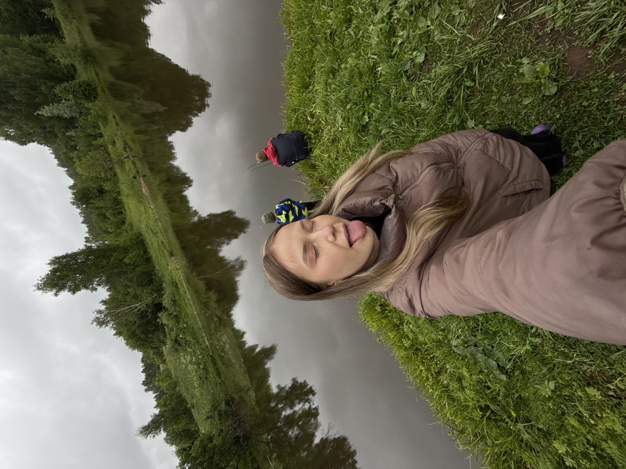 A young woman with long blonde hair wearing a tan jacket is lying on green grass near a body of water. She is sticking out her tongue and has her eyes closed. Two people are visible near the water's edge in the background, one wearing a black and red jacket, the other in a blue and yellow jacket. Both figures appear to be fishing. Trees are visible on the opposite bank of the water. The sky is overcast. The location is likely Zhigalki, Russia.