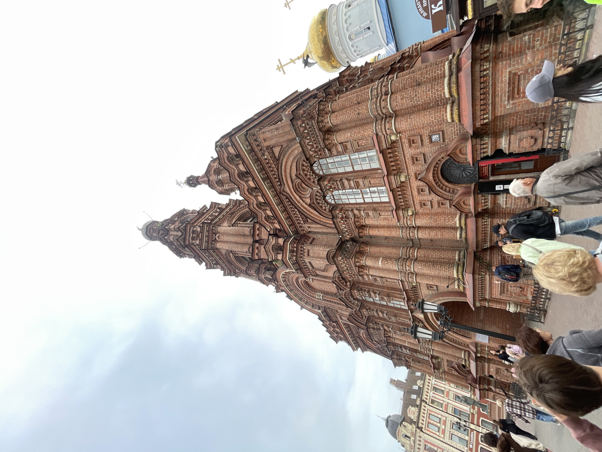 The image depicts a vertical shot of the exterior of the red-brick Peter and Paul Cathedral located in Kazan, Russia. The cathedral features intricate brickwork, ornate detailing, and several prominent domes. A gold-plated dome of a nearby building is partially visible at the top right corner of the image. Pedestrians and tourists are standing and walking on the sidewalk in front of the cathedral. The sky is overcast.