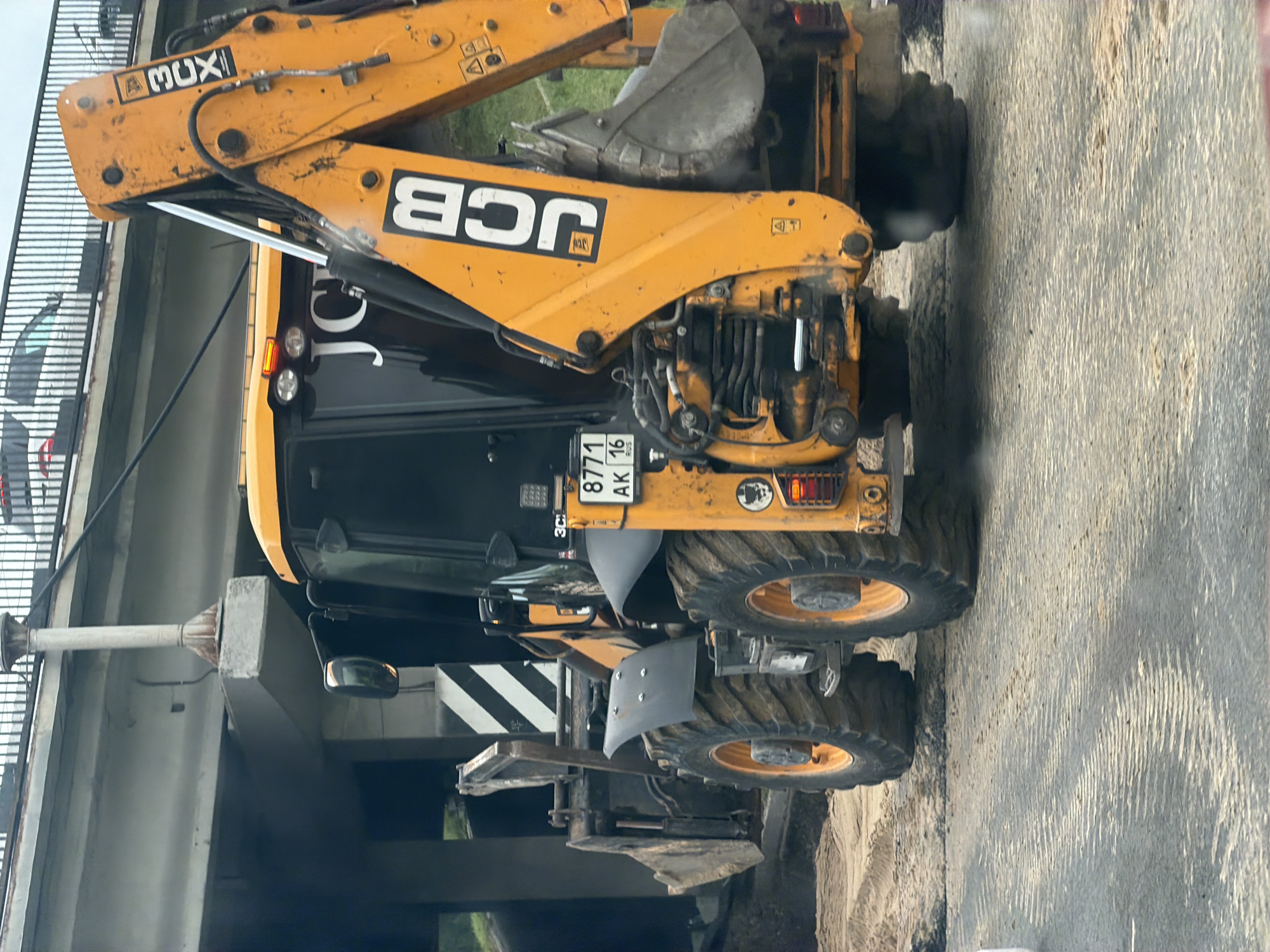 An orange JCB backhoe loader is parked on a sandy surface beneath a concrete overpass, likely a bridge, in Kazan, Russia. The backhoe has a black cabin with large windows, and its name "JCB" is prominently displayed in white letters on its yellow arm. A license plate on the rear of the vehicle reads "8771 AK 16". The ground appears to be a mix of sand and asphalt, with tire tracks visible. The overpass has a metal railing on top, and in the background, the blurry shapes of cars can be seen on what appears to be a road on the overpass. The overall scene suggests ongoing construction or road work.