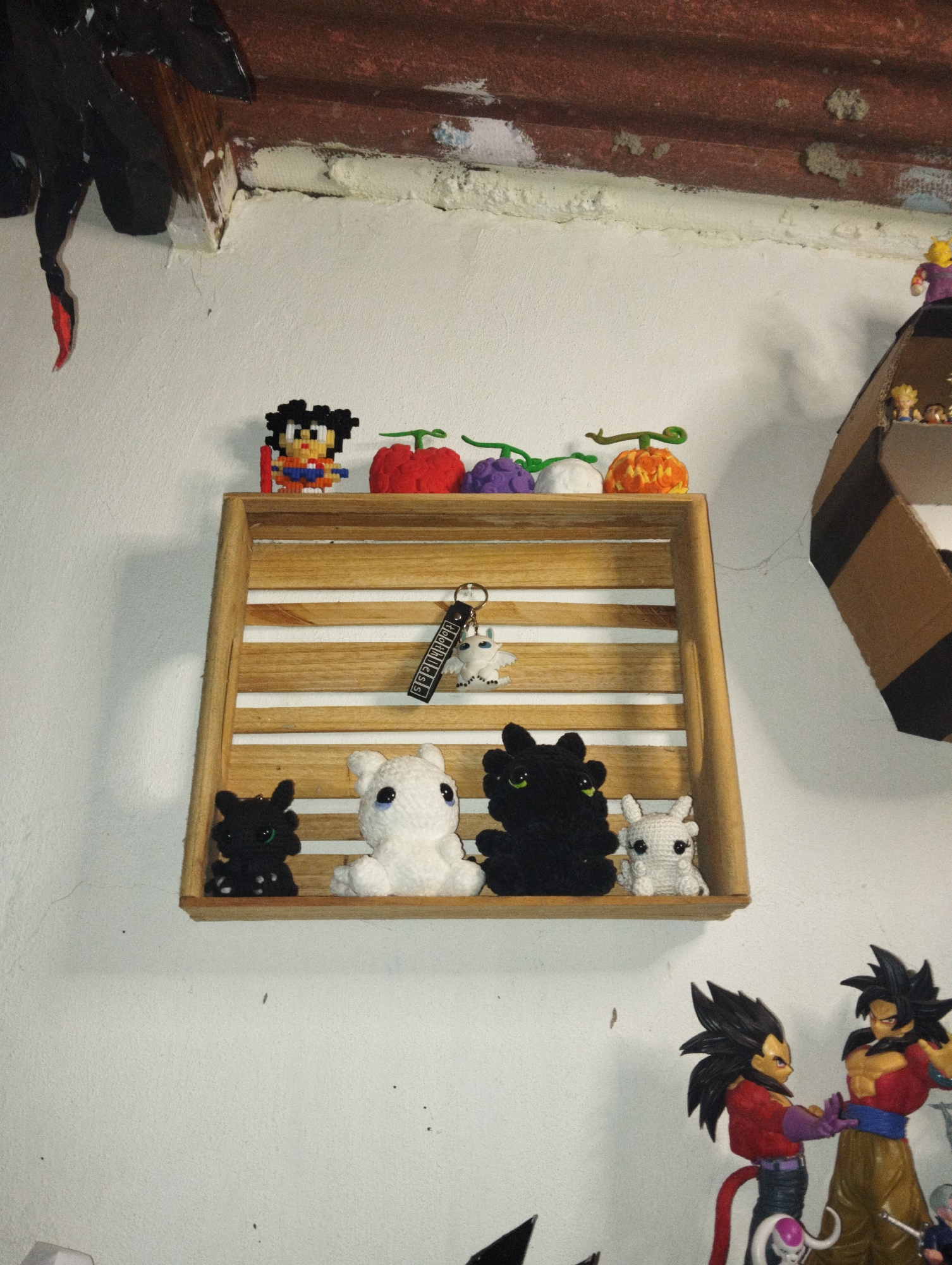 A medium-shot photograph shows a wooden shelf mounted on a white wall. The shelf contains a variety of small figurines and objects, including: pixel art, fruits, a keychain, and crochet figures of Toothless and Light Fury. Below and to the right of the shelf are two Dragon Ball Z figurines. The wall appears to be in a residential interior in El Valle del Espíritu Santo, Venezuela.