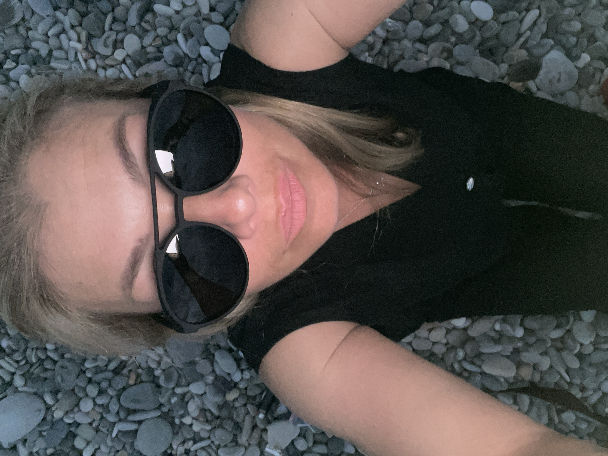 A woman is lying on a pebble beach, wearing large black sunglasses and a black top. She has blonde hair and is looking up at the camera. The pebbles are gray and white and appear to be wet. The lighting is dim, suggesting it may be dusk or early evening.