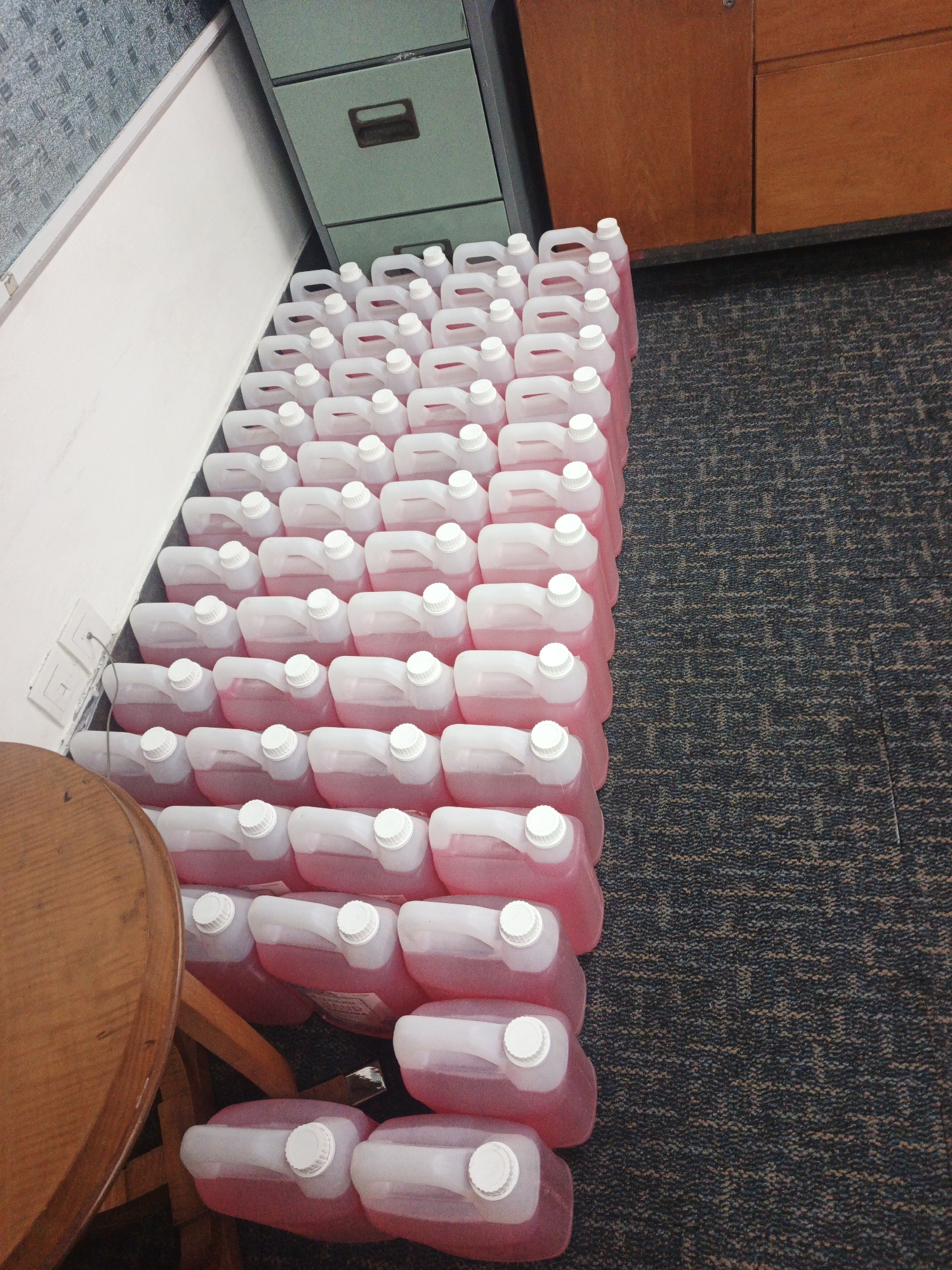The image shows a large quantity of pink liquid in translucent plastic jugs with white caps, neatly stacked and arranged in rows on a carpeted floor. The jugs are lined up against a white wall, next to a filing cabinet and a wooden cabinet. A portion of a round wooden table is visible in the lower left corner. The scene appears to be an indoor storage area. There are no people or animals present. The lighting suggests an indoor environment, possibly an office or storage room. The location is specified as South Jakarta, Indonesia.