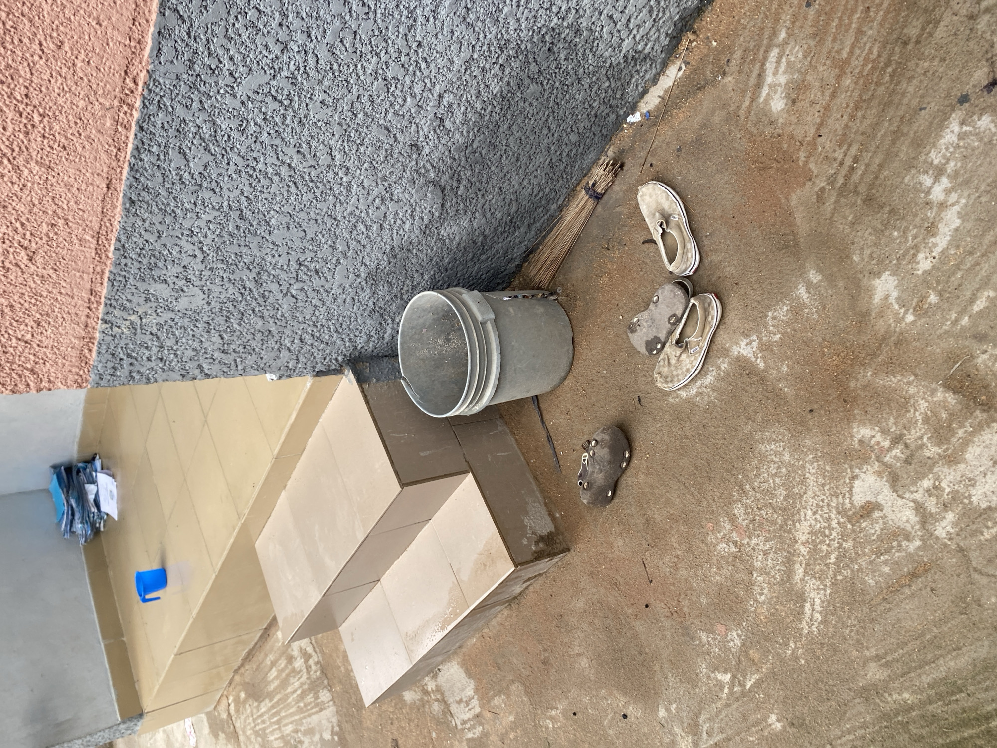 In Jauro Yinu, Nigeria, an outdoor scene depicts a rough, sandy ground with a light-colored tiled step leading to a raised platform. On the platform, a stack of blue-covered papers sits on the tiles. In the foreground, on the ground, are two pairs of worn-out shoes: one pair of dark-colored clogs and one pair of light-colored canvas slip-on shoes. Beside the steps, leaning against a textured gray wall, is a grey plastic bucket and a broom. The wall is painted in two sections: the lower portion is textured gray, and the upper portion is a peachy-pink color.