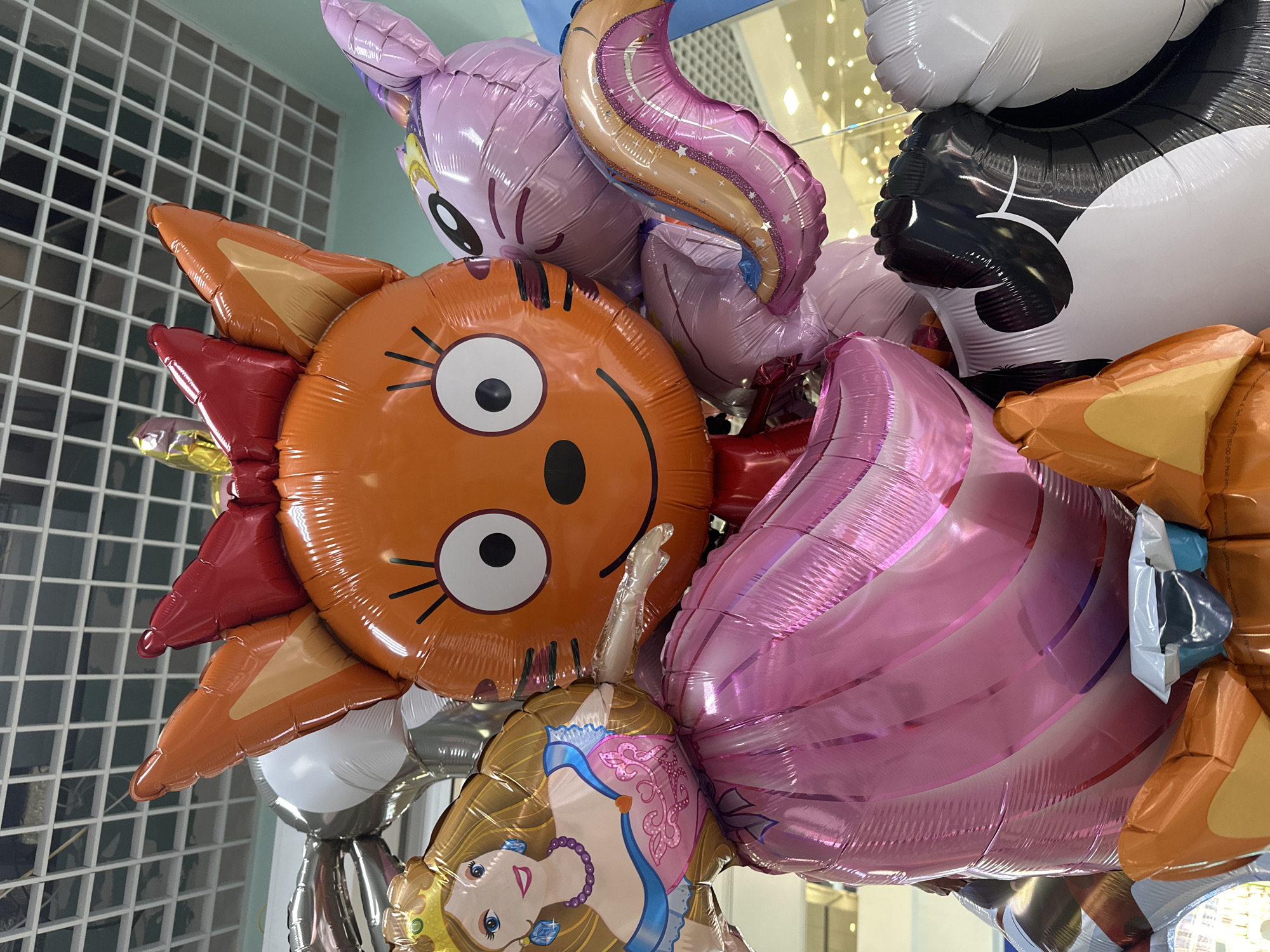A close-up shot shows a cluster of inflated foil balloons. Dominating the foreground is a large orange balloon with a smiling, cartoon-like sun face. It features a round shape, big white eyes, and short black whiskers. Red and orange pointed shapes encircle the face, resembling sun rays.
Above and slightly behind the sun balloon, there is a pink balloon shaped like a pony, with a rainbow-colored horn. Next to the pony, there is a black and white balloon that resembles a cow.
Below the sun balloon, a princess balloon is visible with light skin, blonde hair, and a blue dress. Another pink balloon, possibly shaped like a cupcake or other dessert, is situated to the right of the sun balloon and partially obstructs the view of other balloons.
The background shows a wall with a grid pattern. The top of the scene has a blurred view of what appears to be lights, possibly indicating an indoor location.