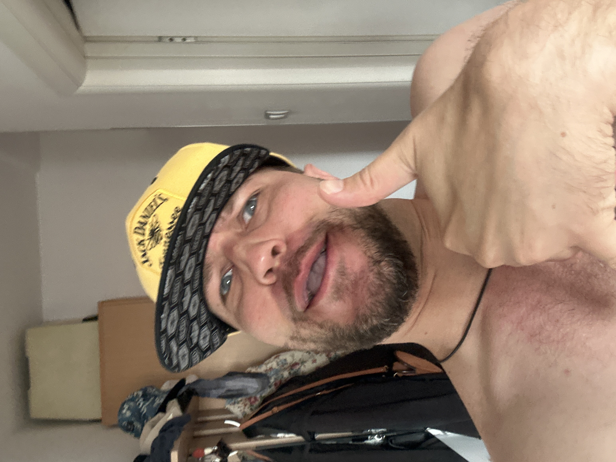 The media file is a selfie taken by a person indoors. The person is wearing a yellow baseball cap with a black and white pattern on the brim, and a black necklace. They have blue eyes and a beard. The person is pointing their index finger towards the camera. The background is blurry but appears to be a room with some furniture. There is visible text on the cap that reads "Jack Daniel's". The image is oriented sideways.