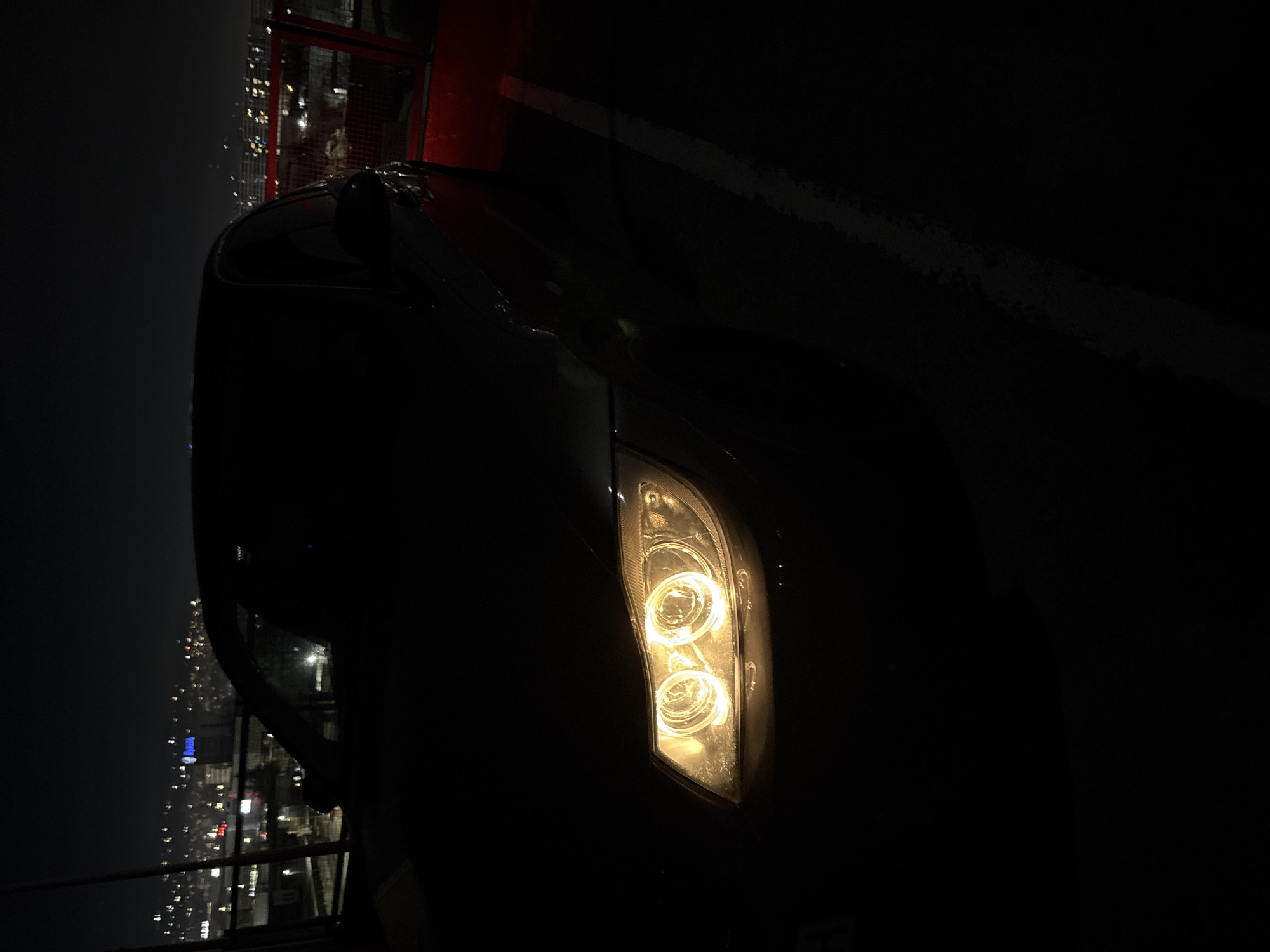The image depicts a night scene, primarily focusing on the illuminated front-right headlight of a car. The car, largely obscured by darkness, appears to be a dark-colored vehicle. The headlight emits a bright, warm yellow light, revealing its internal structure with two distinct circular light sources.
In the background, a blurry urban landscape stretches out, characterized by numerous scattered lights from buildings and streets, suggesting a cityscape at night. The perspective implies the car is situated at an elevated position, possibly overlooking the city, or within a structure that allows a view of the city lights. To the left of the car, a metal railing or fence is visible, further hinting at an elevated location like a bridge, parking garage, or high viewpoint. A red structure, possibly part of a building or a large sign, is also faintly discernible in the upper left section of the background.
The overall time of day is clearly night, with darkness dominating the scene, punctuated only by the car's headlight and the distant city lights. No specific activity or people are visible, and no text is discernible. The setting is consistent with an urban environment like Varna, Bulgaria, as provided by the context.