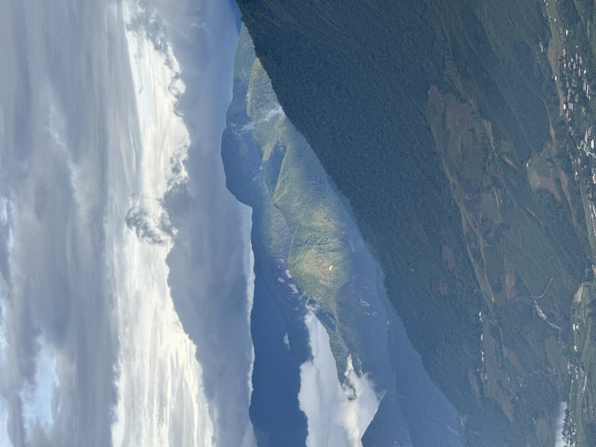 The image depicts a mountainous landscape in Sa Pa Ward, Vietnam, with a layered vista of ridges covered in dense green vegetation. Patches of cultivated land are visible on the slopes. The sky is filled with dramatic cloud formations, ranging from bright white to dark grey. A diffused sunlight illuminates a portion of one of the mountain ridges. The image is oriented in a vertical format.
